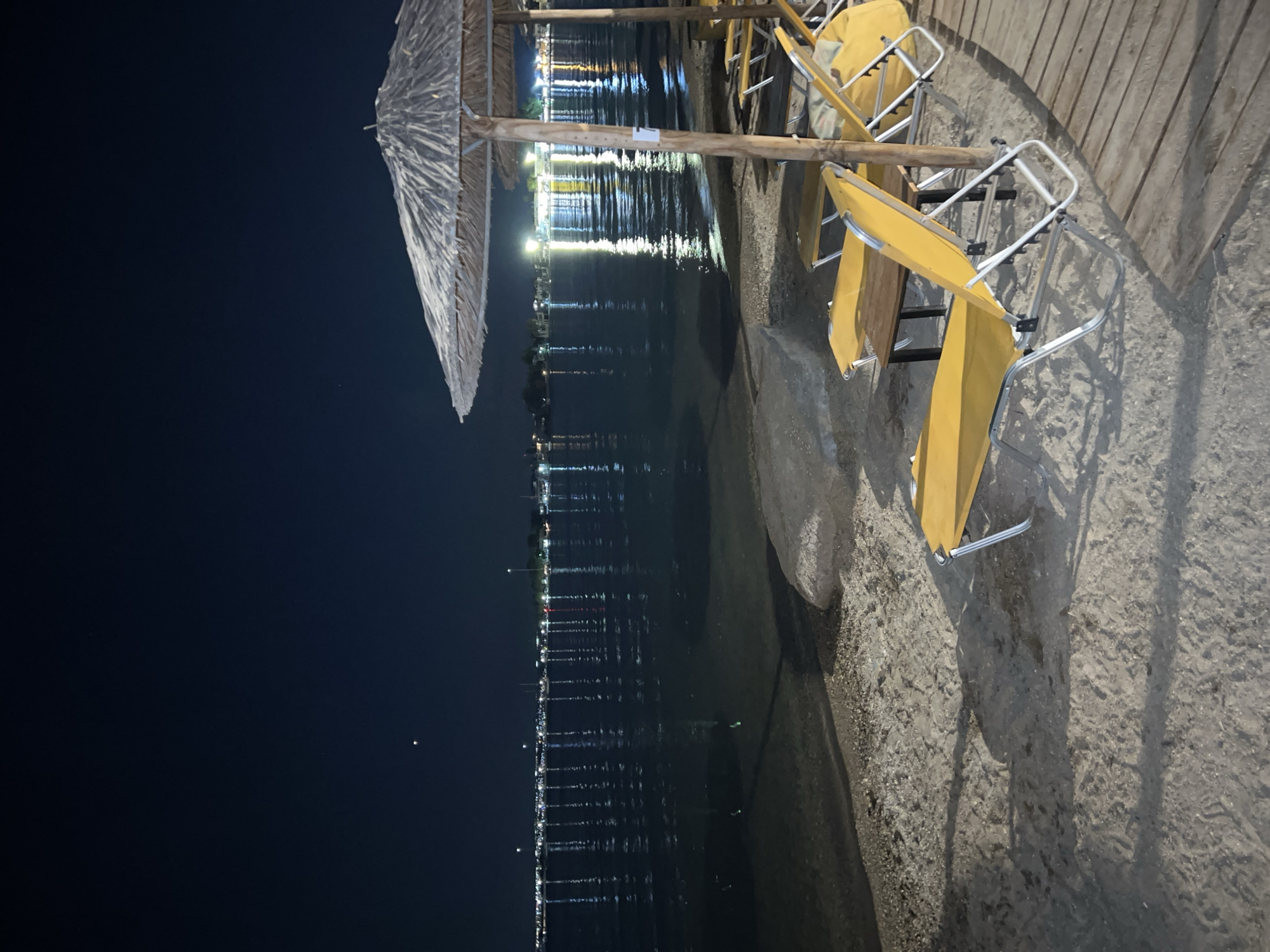 The image depicts a beach at night in Eretria Municipal Unit, Greece, facing a body of water with distant lights reflecting on its surface. The foreground features several empty yellow lounge chairs arranged on the sand, some partially covered by a straw umbrella. A small wooden table is visible between two of the chairs. The wooden deck runs along the right side of the frame, leading into the sand. The water is calm, with noticeable reflections of the lights from the far shore. The sky is dark blue, with a single bright star or celestial body visible. There are no people present in the scene.