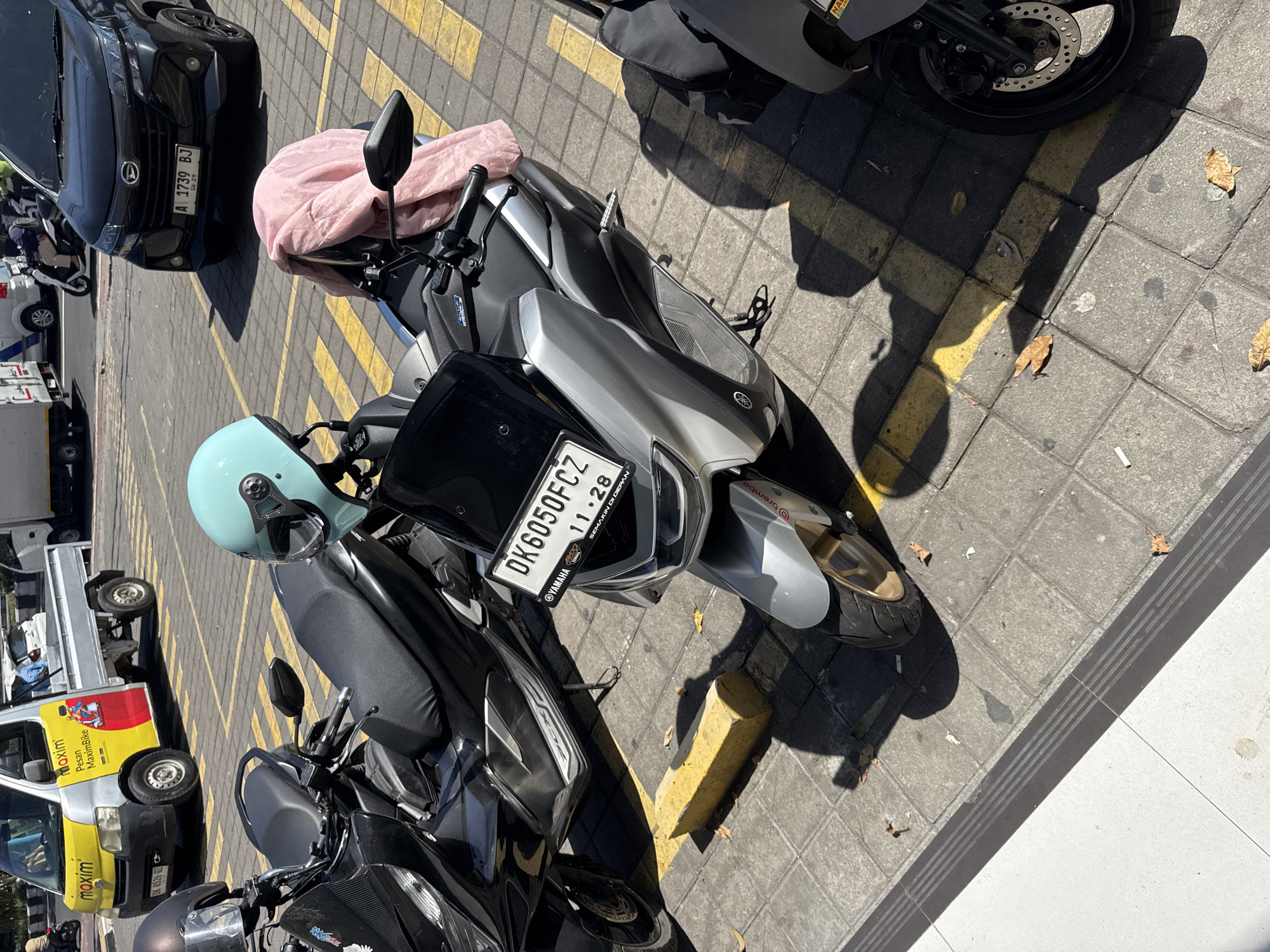 In Kuta, Indonesia, parked motorcycles occupy a sunlit area paved with grey interlocking bricks, marked with yellow lines. A light blue motorcycle helmet rests on a black motorcycle, which is parked next to a silver motorcycle displaying a license plate reading "DK6050FCZ" and "11.28". Another black motorcycle is visible behind the silver one. In the background, a white pickup truck with yellow and red branding for "maxim Pesan MaximBike" is parked, with a dark SUV to its right. The scene suggests a public parking area in Kuta.