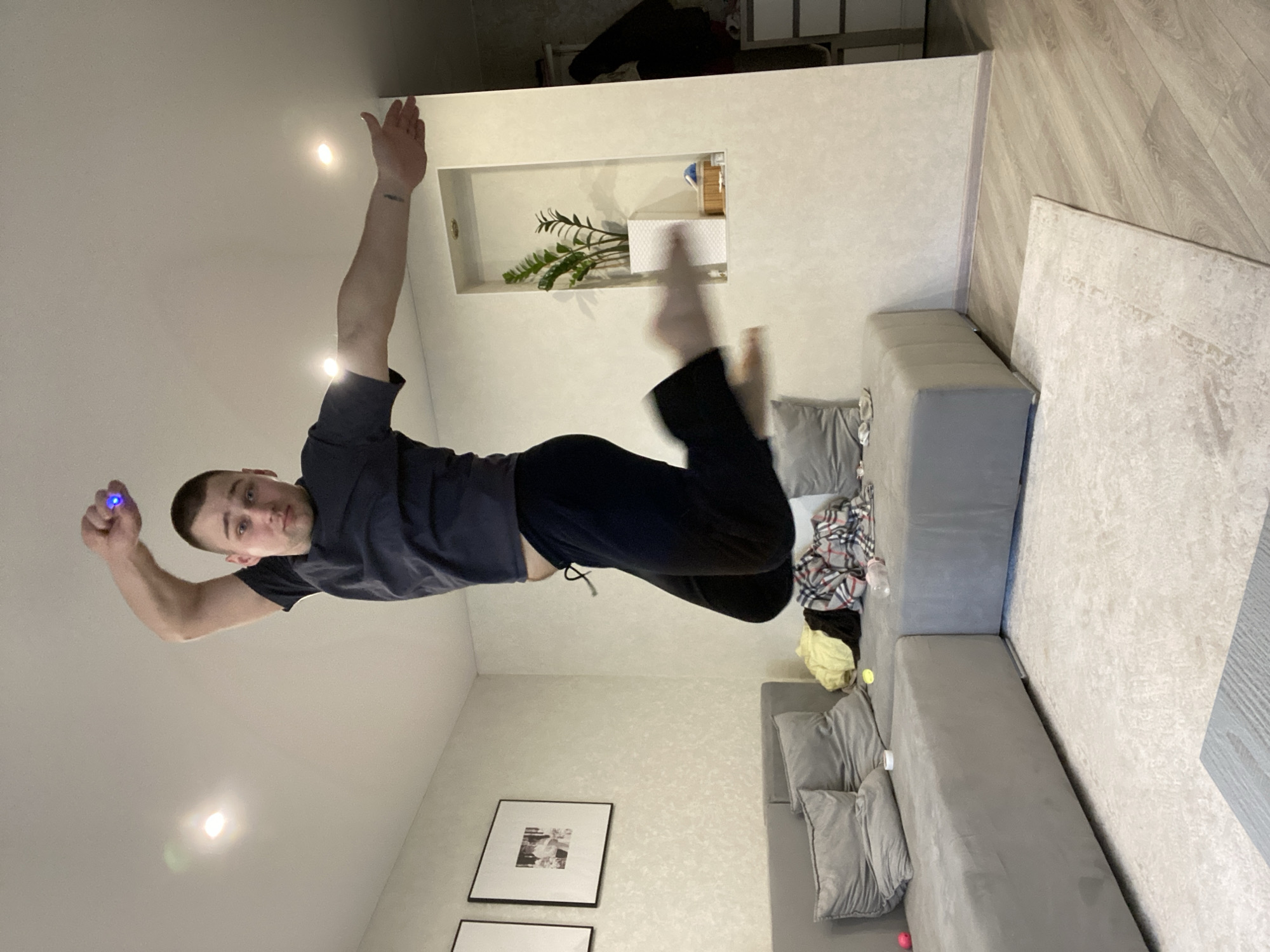 The scene is an interior view of a residential living space, likely an apartment in Chaikovsky, Russia. A male individual, mid-air, is captured in the center of the frame, appearing to jump. He has short dark hair, is fair-skinned, and is wearing a dark short-sleeved t-shirt and dark athletic pants. His right arm is raised with the hand clenched, holding a small object emitting a blue light. His left arm is extended outwards, palm open, revealing a small dark tattoo on his forearm. His legs are bent, and his feet are bare.
The room features light-colored, possibly textured, walls. The ceiling is white with multiple recessed spotlights, two of which are visible and illuminated. A large, grey L-shaped sectional sofa occupies the lower-left and central portions of the image, adorned with several grey pillows. Assorted clothing items, including a yellow garment, a plaid fabric, and a dark item, are resting on the right section of the sofa, along with a clear plastic bottle and a small pink ball.
The floor consists of a light-colored, patterned rug covering the central area, with light-toned laminate wood flooring visible around its perimeter. On the wall above the left section of the sofa, two framed pictures with black frames and white matting are displayed. To the right, a recessed wall niche contains a green potted plant in a white textured planter, alongside a small wooden box and other minor items. Further to the right in the background, a white cabinet with drawers and a dark item of clothing draped over it are partially visible.