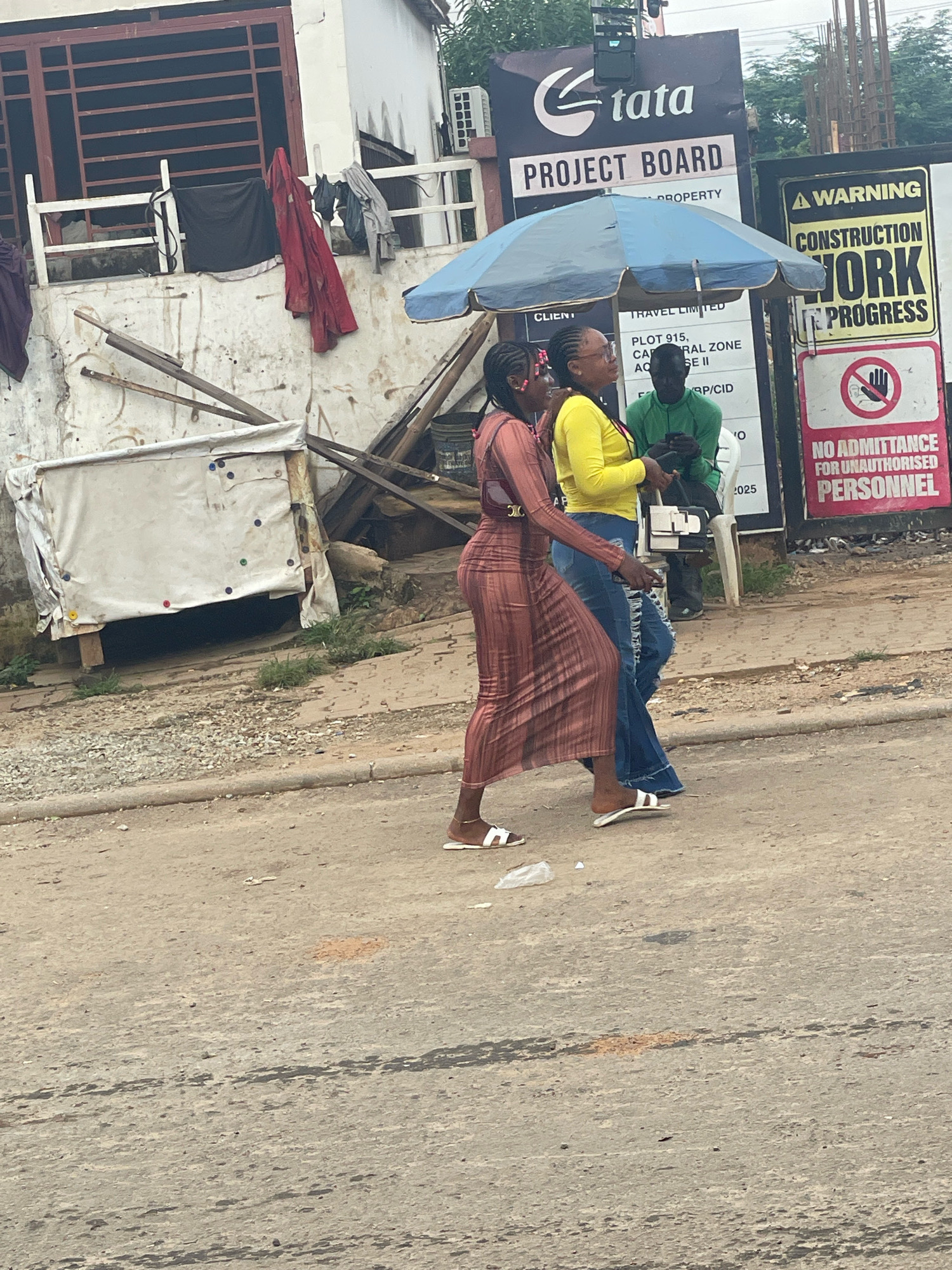 In Abuja, Nigeria, two women are walking on a dirt road. One woman, on the left, is wearing a long, brown, patterned dress and white sandals. The other woman, to her right, is wearing a bright yellow top and ripped blue jeans, also with white sandals. Behind them, a man in a green shirt is looking at his phone. A blue umbrella provides shade for a group of people. In the background, a building with a white exterior has clothes hanging on a railing. A sign for "Tata Project Board" is visible, as well as a "Warning: Construction Work in Progress" sign with a "No Admittance for Unauthorized Personnel" notice. The environment appears to be an urban street with construction activity and some signs of wear and tear on the road and buildings.