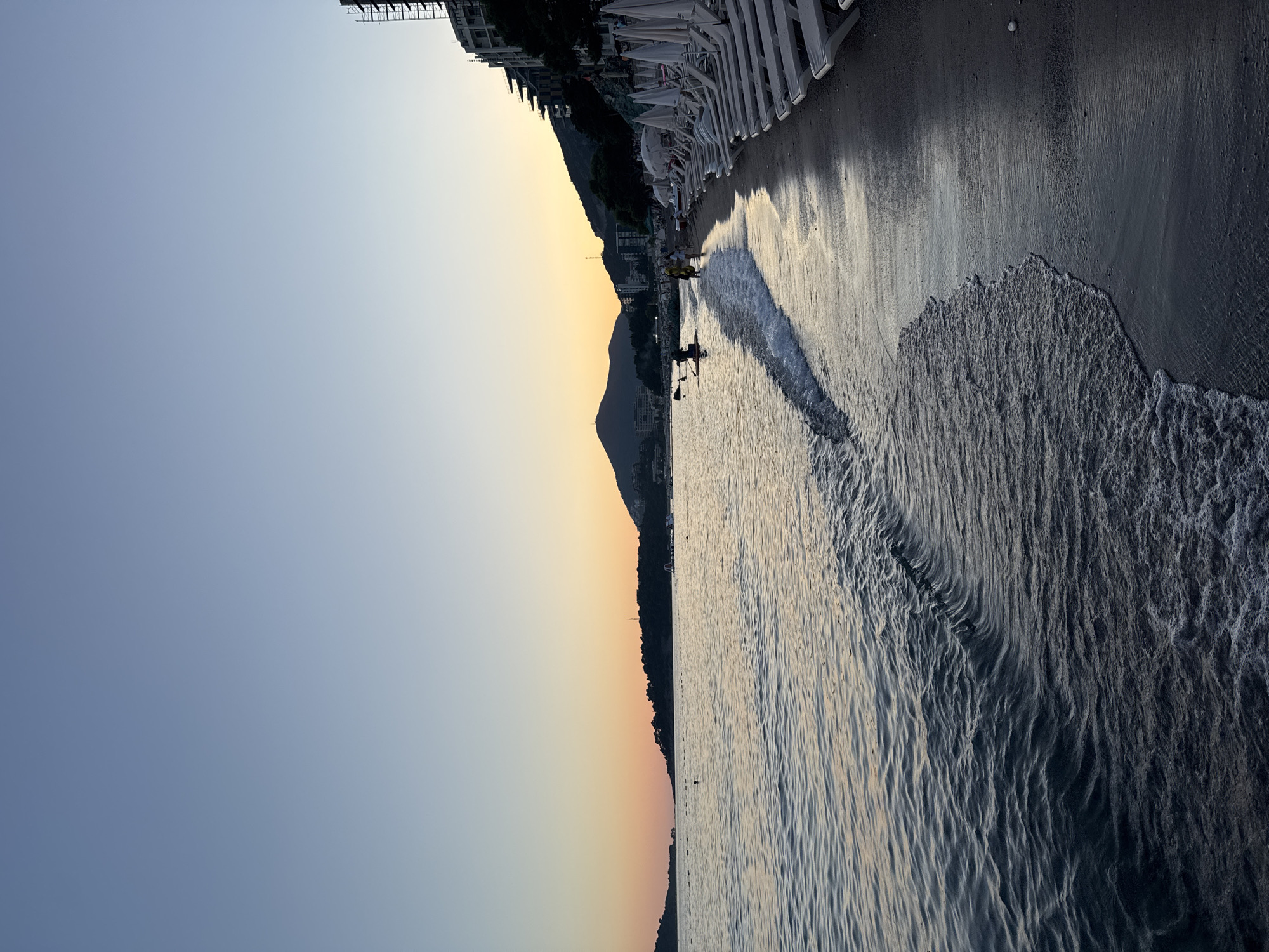 The image depicts a serene beach scene in Rafailovići, Montenegro, during the soft light of dusk. The sea gently laps at the shore, with small waves creating a foamy edge along the dark sand. The sky transitions from a clear, pale blue at the top to a warm gradient of orange and pink near the horizon, suggesting the sun has recently set or is about to rise.
In the foreground, the dark, wet sand reflects the muted colors of the sky. A line of empty, white lounge chairs and umbrellas is neatly arranged along the edge of the beach, ready for the next day. Further down the beach, a few figures are visible. One person is on a paddleboard in the water, their silhouette dark against the lighter sea. Two people are walking along the shoreline, one appearing to be wearing a yellow garment.
The background features a hilly landscape covered with dark vegetation and dotted with buildings, indicative of a coastal town. A tall, unfinished building with scaffolding is prominent on the right side of the frame, contrasting with the natural beauty of the scene. The overall atmosphere is tranquil and peaceful, capturing the quiet end of a day at the beach.