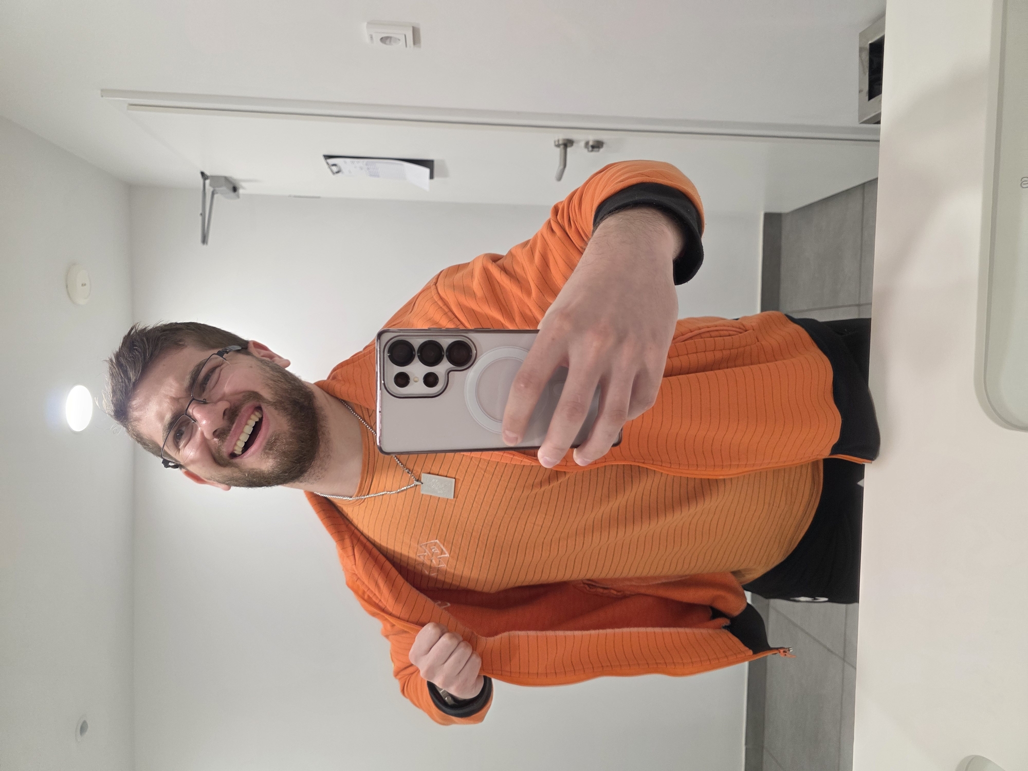 A man with glasses and a beard is taking a selfie in what appears to be a bathroom. He is wearing an orange shirt and jacket, and holding a smartphone with multiple camera lenses. The background shows a white door, wall tiles, and bathroom fixtures, suggesting the location is inside a building. The specific location in Plovdiv, Bulgaria is not discernible from the image.