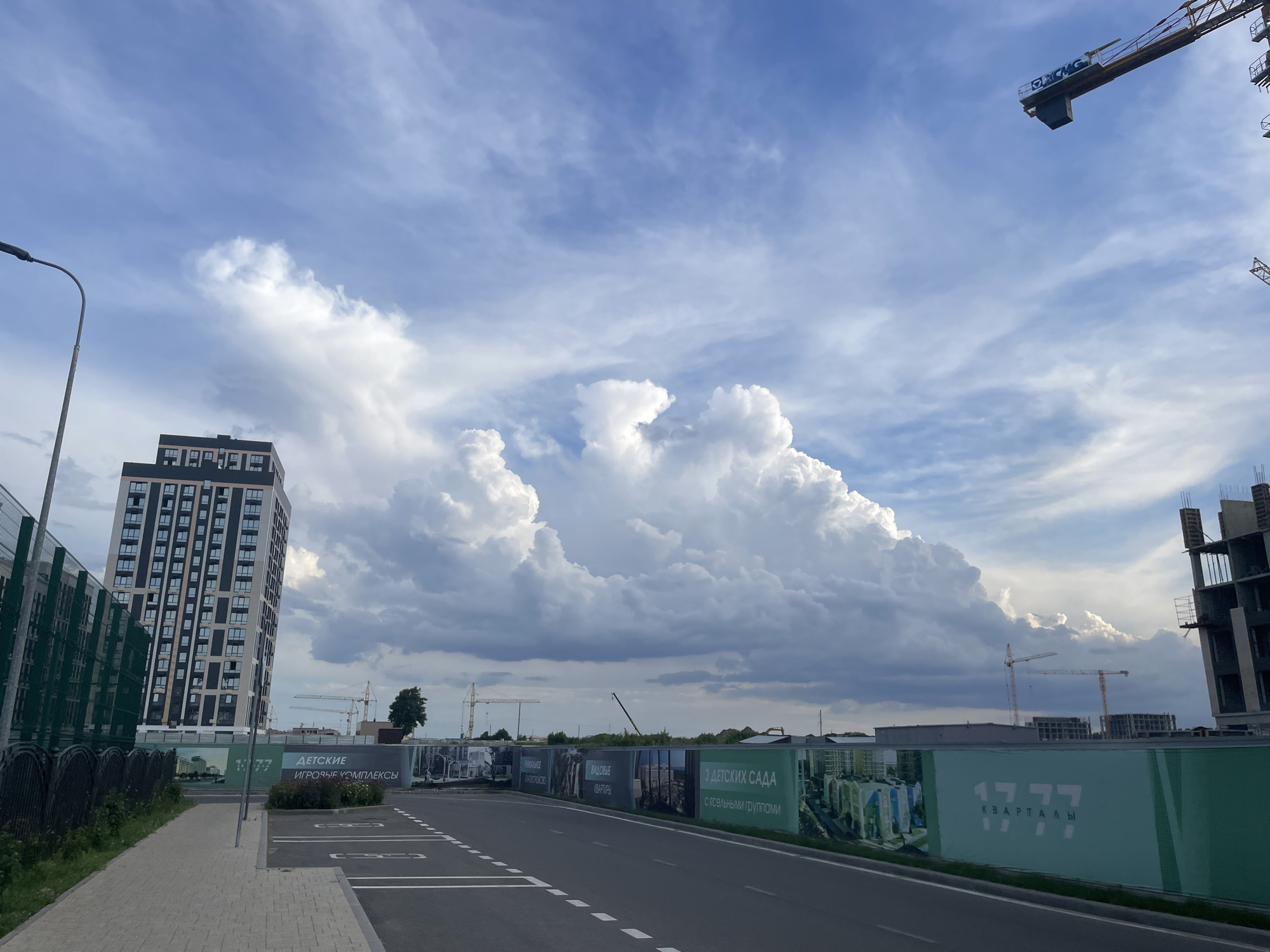 The image depicts a construction site in the city of Stavropol, Russia, under a partly cloudy sky. In the foreground, a paved road with white dashed lines runs horizontally, leading towards a sidewalk. To the left of the road is a sidewalk made of light-colored paving stones, bordered by a green fence. A tall, modern apartment building with numerous windows stands prominently on the left side of the image.
In the background, several construction cranes are visible, indicating ongoing development. A large banner with green and white graphics and Russian text, likely advertising residential properties, stretches along a fence. The banner features the number "17 77" and the word "КВАРТАЛЫ" (quarters/blocks). To the right of the banner, unfinished multi-story buildings are under construction. The sky is a mix of blue and white clouds, with a large, dramatic cloud formation dominating the center of the frame. The overall scene suggests a developing urban area.