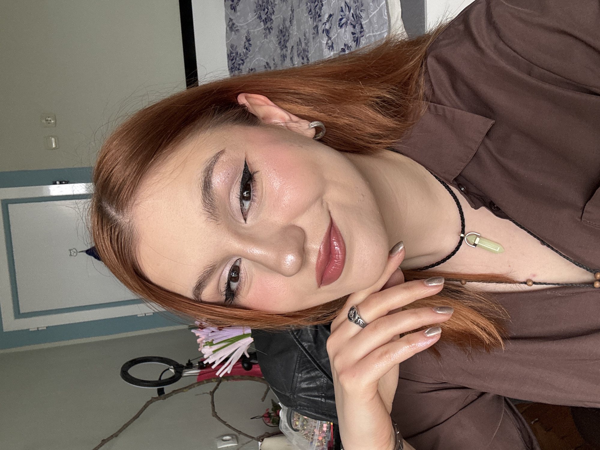 A person with fair skin, reddish-brown hair, and makeup including winged eyeliner, blush, and lip color poses for a photograph. The individual wears a brown collared shirt and a black cord necklace with a light green pendant. Their left hand, adorned with a ring and metallic gray nail polish, is positioned near their face. The background includes a white-framed wall, a patterned curtain, a black leather jacket, a ring light, and branches with flowers. There is no information available in the image to suggest it was taken in Varna, Bulgaria.