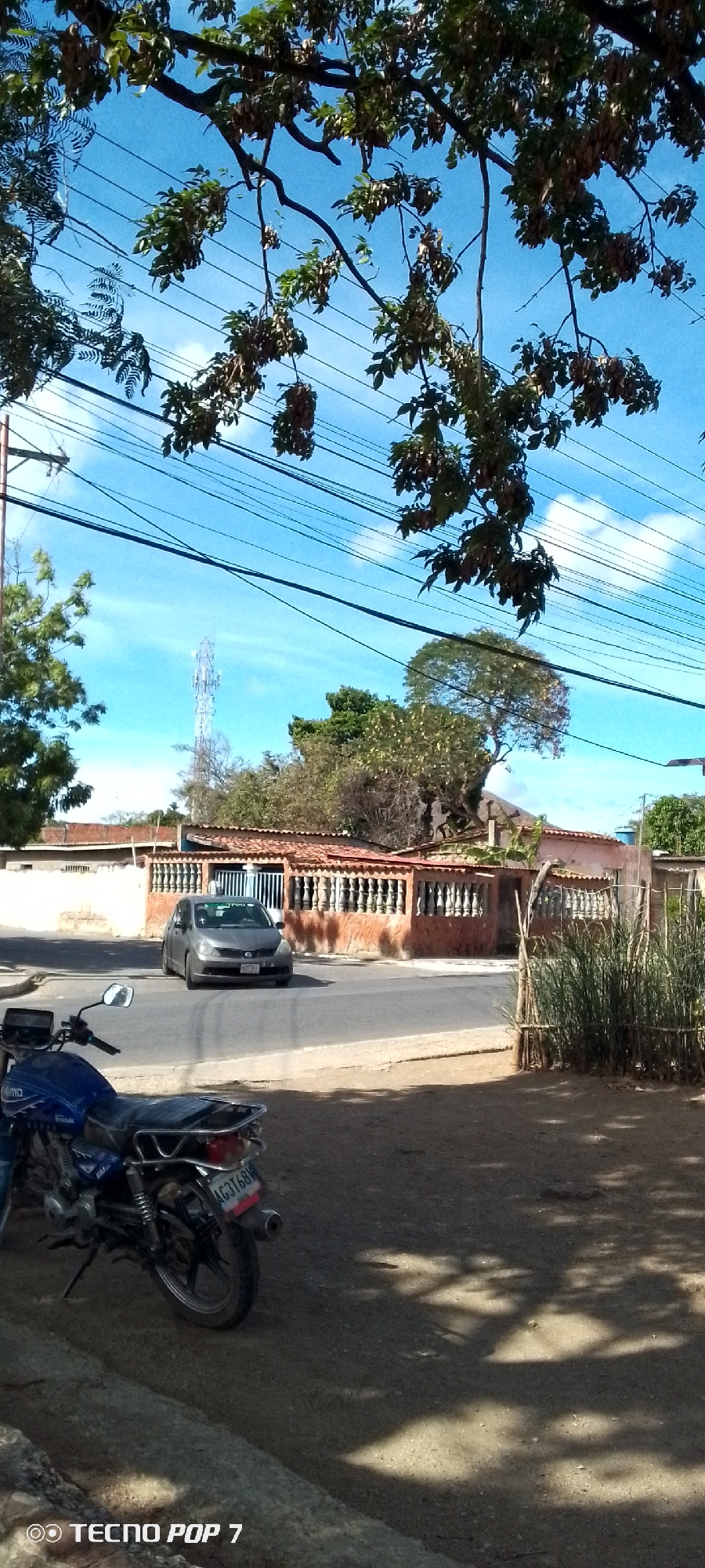 The image shows a street scene in Altagracia, Venezuela. A blue motorcycle is parked in the foreground. A grey car is driving on the street. A building with a red tile roof is located along the street. Overhead power lines cross the sky. Trees with foliage are present in the scene. A communication tower is visible in the background. The sky is blue with some clouds.