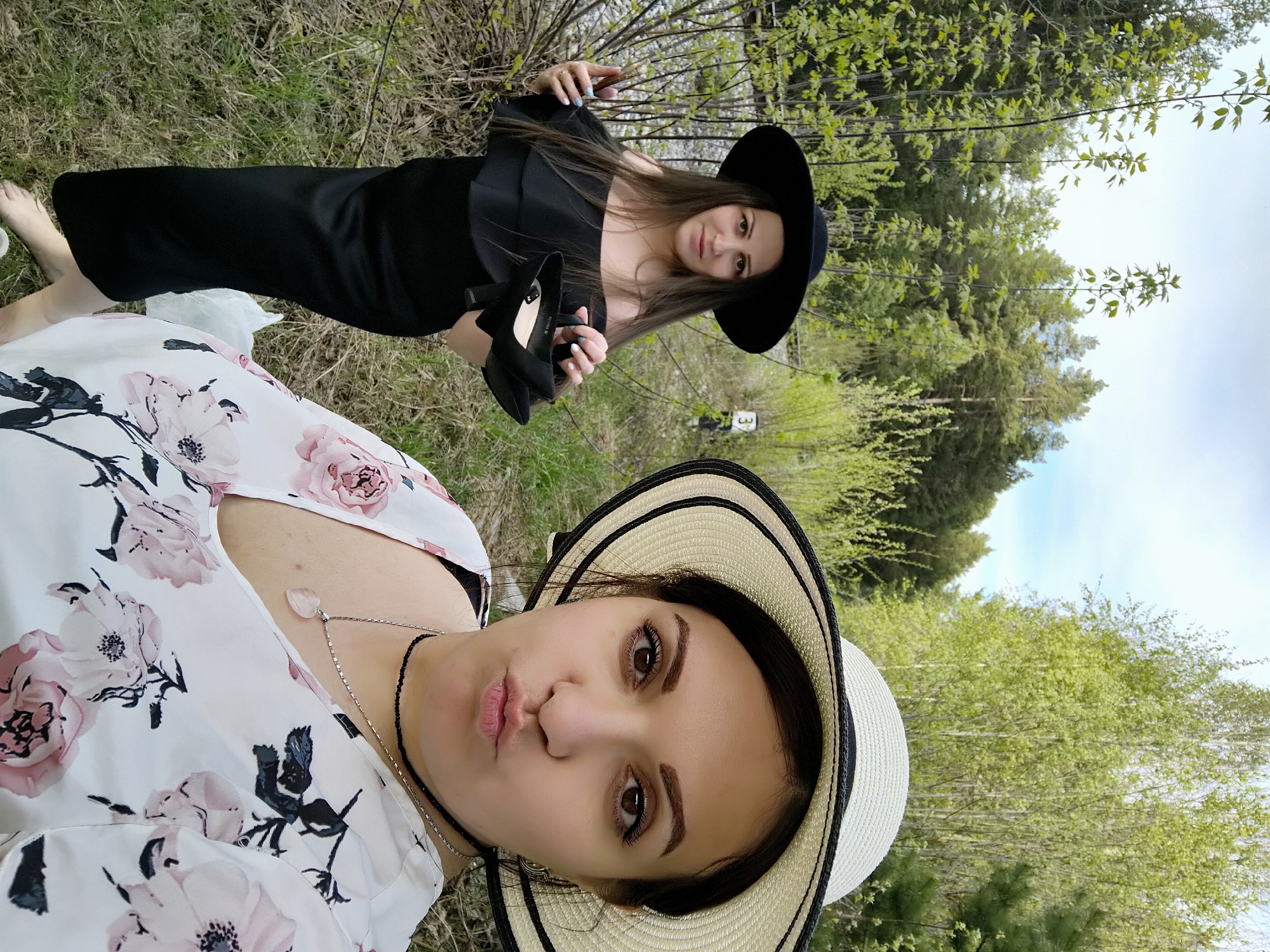 The image depicts two women outdoors, presumably in Chaikovsky, Russia, given the prompt. The foreground features a woman in a floral-patterned dress and a straw hat with a black band, posing with a pout. Behind her, partially obscured by foliage, is another woman wearing a black dress and a wide-brimmed black hat, holding black high-heeled shoes in her hands. The background consists of trees and a glimpse of the sky. The environment appears to be a park or wooded area. The women seem to be interacting or posing for a photograph together.