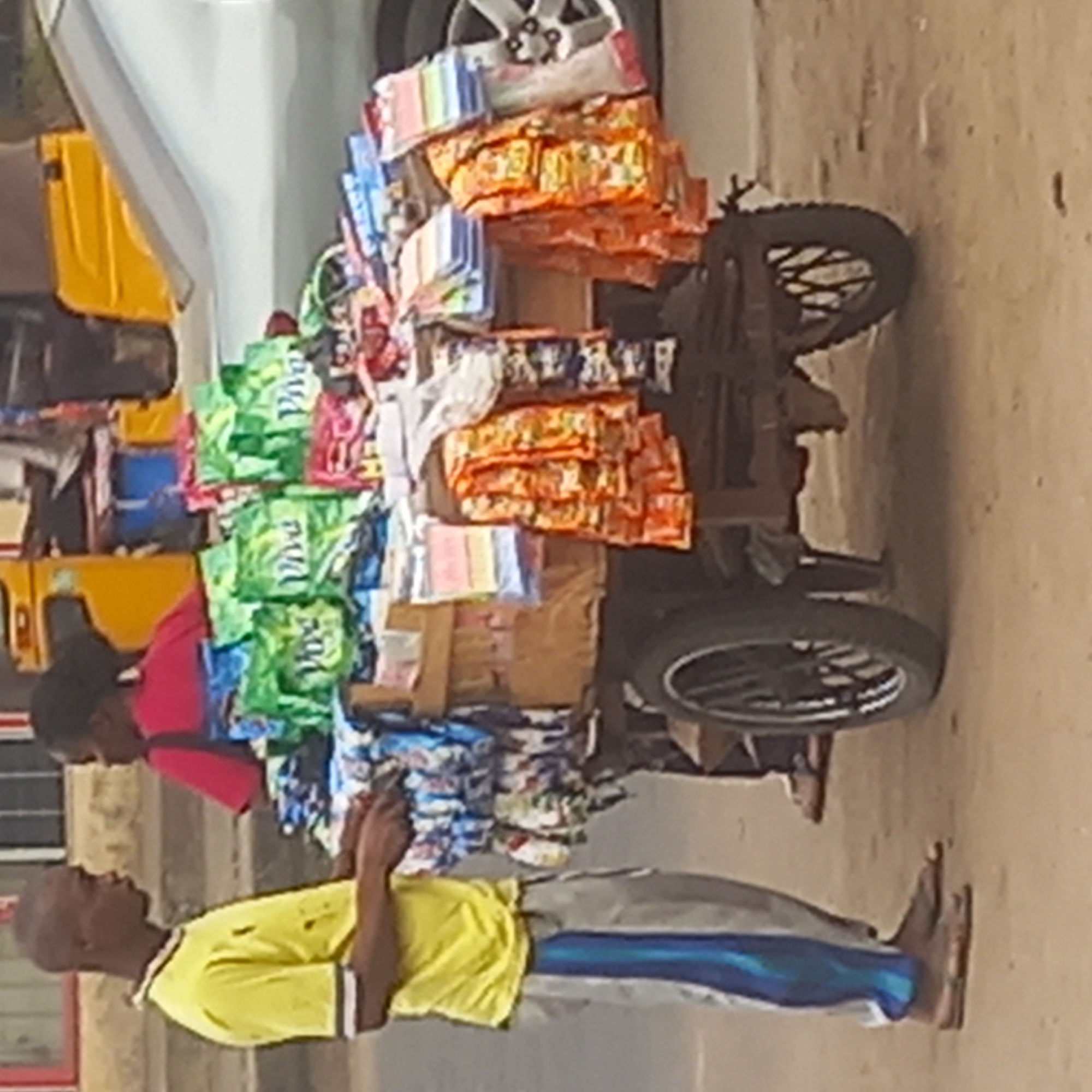 A street vendor stands in Dadi Riba, Nigeria, wearing a yellow t-shirt and grey pants with a blue stripe. He's holding a mobile phone. Next to him, another man in a red shirt stands near a three-wheeled motorcycle cart. The cart is heavily loaded with various packaged goods, including detergent and snacks, stacked high. A car is partially visible on the left of the cart. The setting appears to be an urban street.
