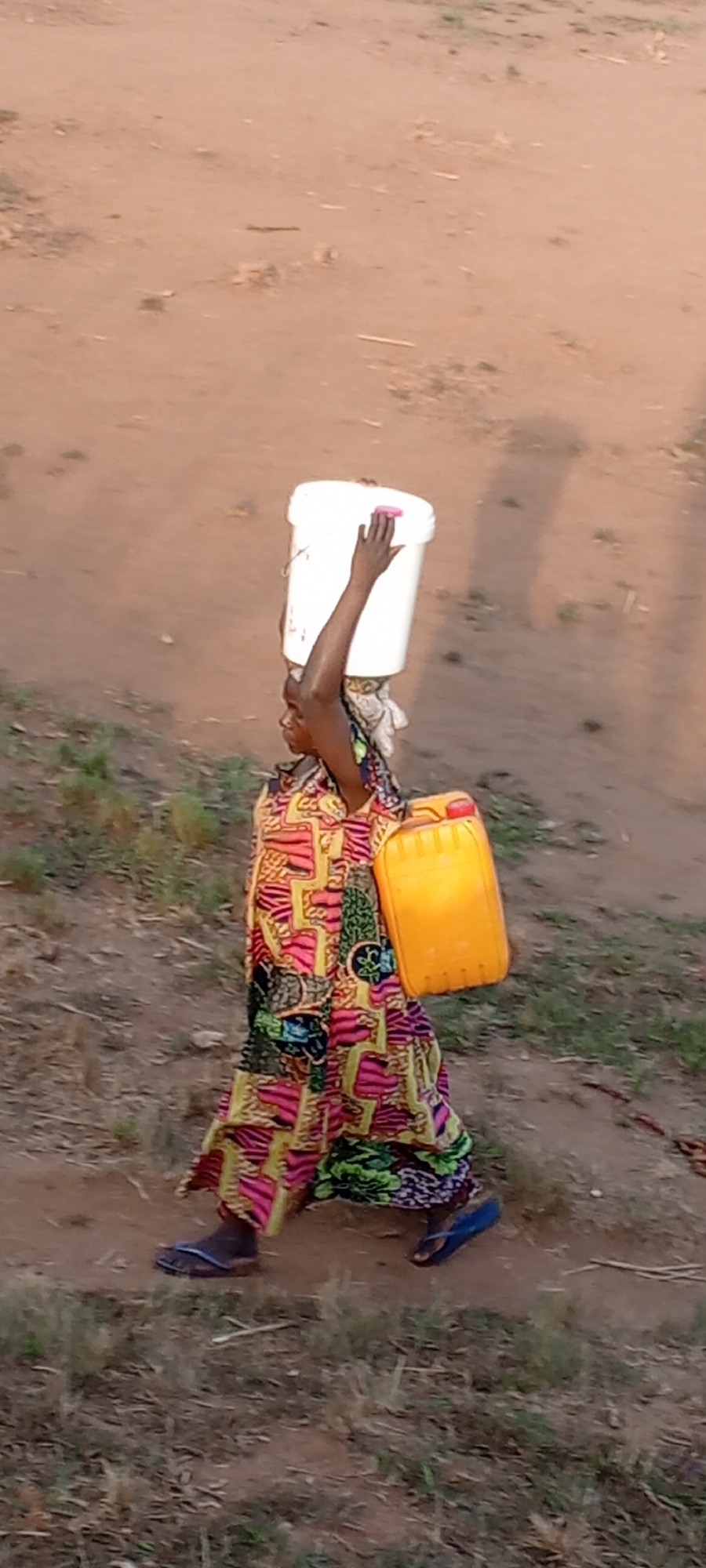 A woman is depicted walking along a dry, reddish-brown dirt path in Ngalula, Democratic Republic of the Congo. She is shown in profile, facing towards the left side of the frame. On her head, she carries a large white bucket, steadying it with her right arm raised and hand placed on the bucket's rim. A small pink object is visible on top of the white bucket. In her left hand, she carries a bright yellow jerrycan close to her side.
The woman is wearing a long, loose-fitting dress adorned with vibrant, multi-colored patterns, primarily featuring shades of pink, green, yellow, and black. She has blue flip-flops on her feet. The ground is largely barren earth with scattered patches of dry grass and small plants. Distinct shadows, including those of the woman and the containers she carries, are cast on the ground, stretching to the right, indicating the photograph was taken during either early morning or late afternoon under clear, sunny weather conditions. The activity suggests the daily chore of transporting water or other liquids in a rural setting. No text is visible in the image.