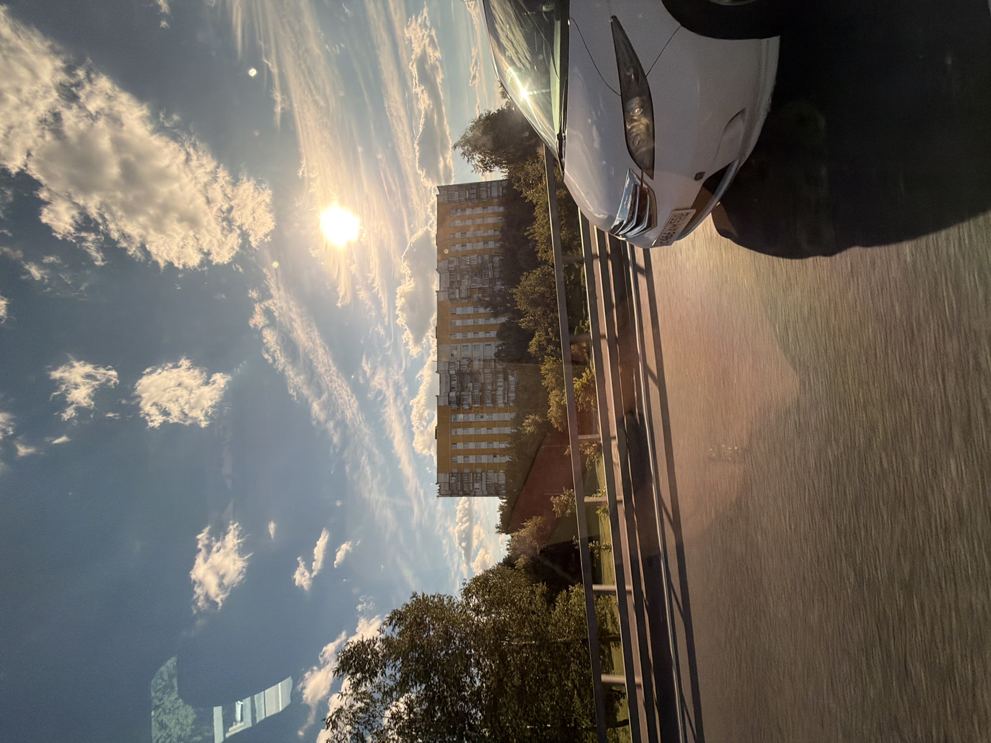 In the city of Moskovskiy, Russia, a white minivan is captured in motion on a sun-drenched road. The vehicle is in the foreground, partially obscuring a long, yellow apartment building in the background. The building is surrounded by trees and appears to be situated in a residential area. The sky above is a vivid blue, dotted with white, fluffy clouds and illuminated by a bright sun. The lighting suggests it is late afternoon or early evening, casting long shadows across the road and the side of the minivan. A metal railing runs along the side of the road, separating it from a grassy area below. The overall scene depicts a typical urban landscape bathed in the warm glow of the setting sun.