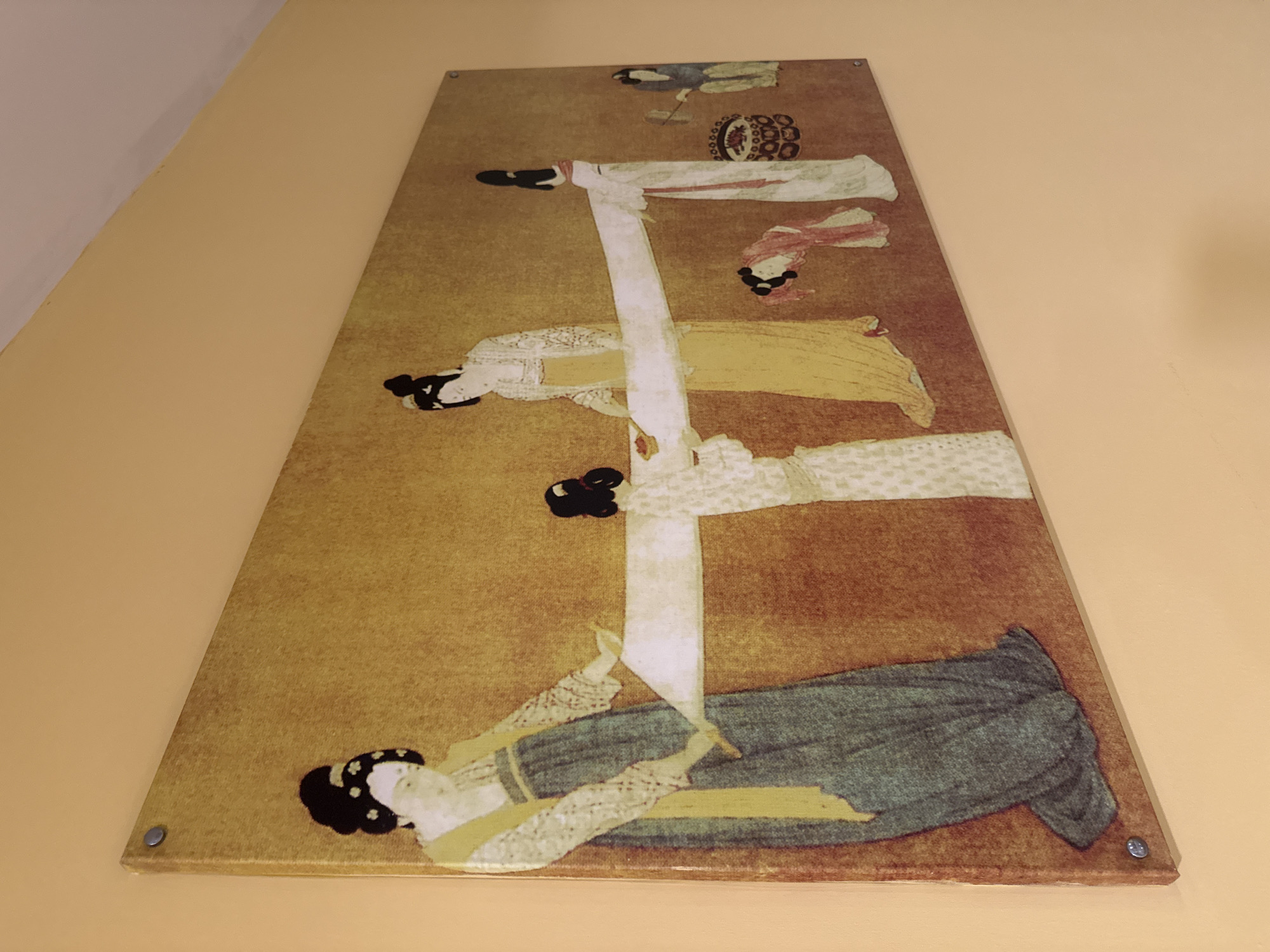 This is a photograph of a rectangular artwork mounted on a light peach-colored wall. The artwork depicts a scene with several figures in traditional East Asian attire, rendered in a flat, illustrative style with a muted color palette. The background of the artwork is a textured golden-yellow. The figures appear to be engaged in an activity involving a long piece of white fabric stretched between them.
The leftmost figure, a woman with her hair in an updo adorned with small white flowers, wears a white patterned robe over a yellow undergarment and a long, dark blue skirt. She is holding the white fabric with her left hand. To her right, a figure in a white patterned robe and yellow skirt stands, also holding the fabric. Further to the right, a figure in white with a red sash holds the fabric. There is a smaller figure, possibly a child, kneeling and looking at the fabric. On the far right, a figure in a blue garment appears to be working with a basket and a tool.
The artwork is mounted on the wall using visible silver screws in the corners. The lighting in the room is warm, casting subtle shadows. The photograph is taken from an angle, showing the side of the artwork and its slight protrusion from the wall.
This scene is not located in Aksu, Turkey. The artwork's style and subject matter suggest an East Asian origin, possibly Chinese.