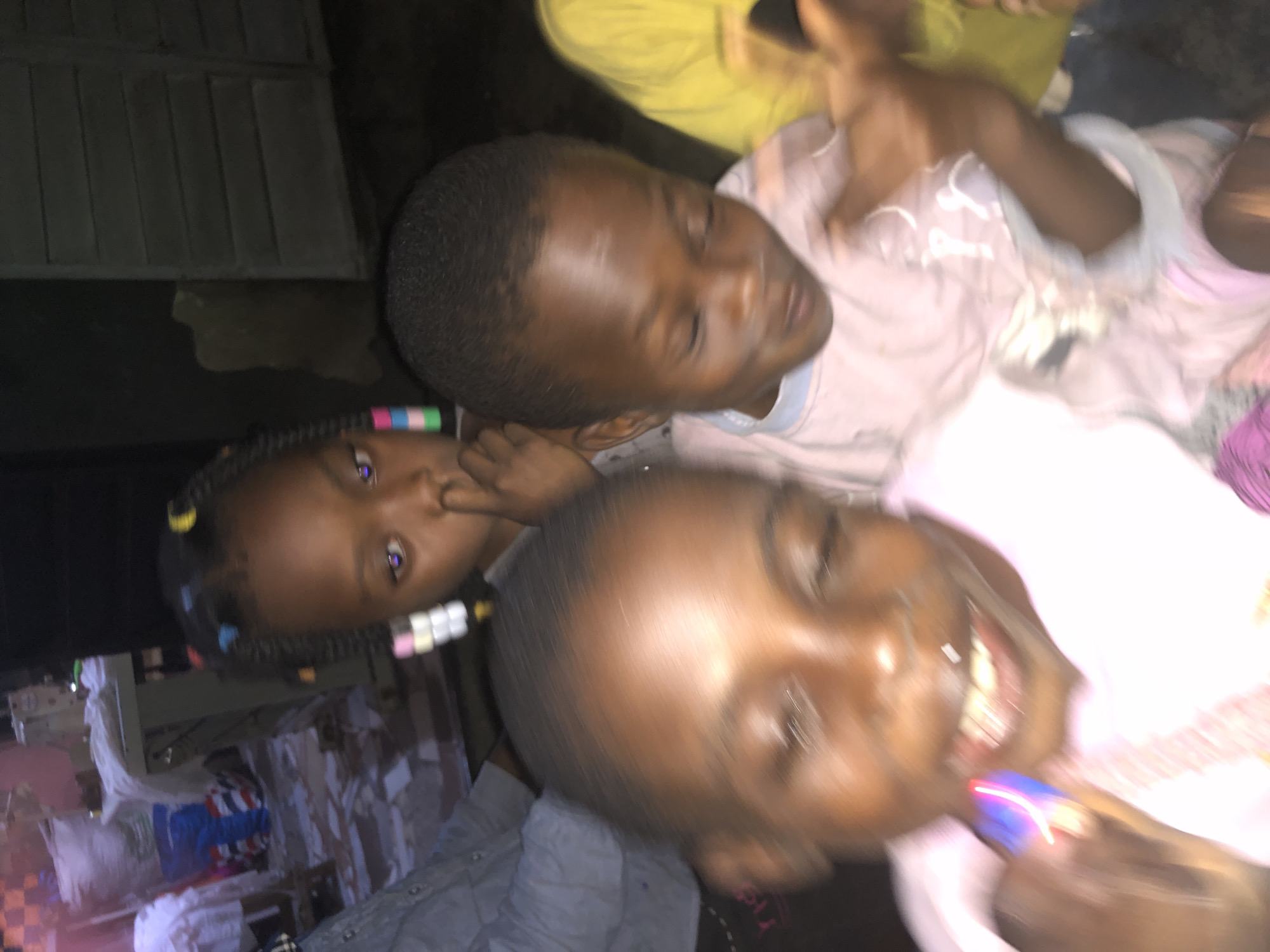 In Ado, Nigeria, a group of three young children are the focus of this dimly lit, candid photograph. The child in the foreground on the left is blurry and appears to be holding a small, colorful object, possibly a toy, near their mouth. Their eyes are open and looking forward. To the right of this child, another child's face is visible, also blurry, with their eyes closed and head tilted down. Behind them, a third child is positioned with their face turned towards the camera, their left eye visible, and their right hand obscuring part of their mouth. This child has braids with colorful beads. In the background, hints of a workshop or storage area are discernible, with large white bags, fabric, and a sewing machine visible. The overall impression is of a casual, indoor gathering, possibly a home or community space, at dusk or night, given the lighting conditions.
