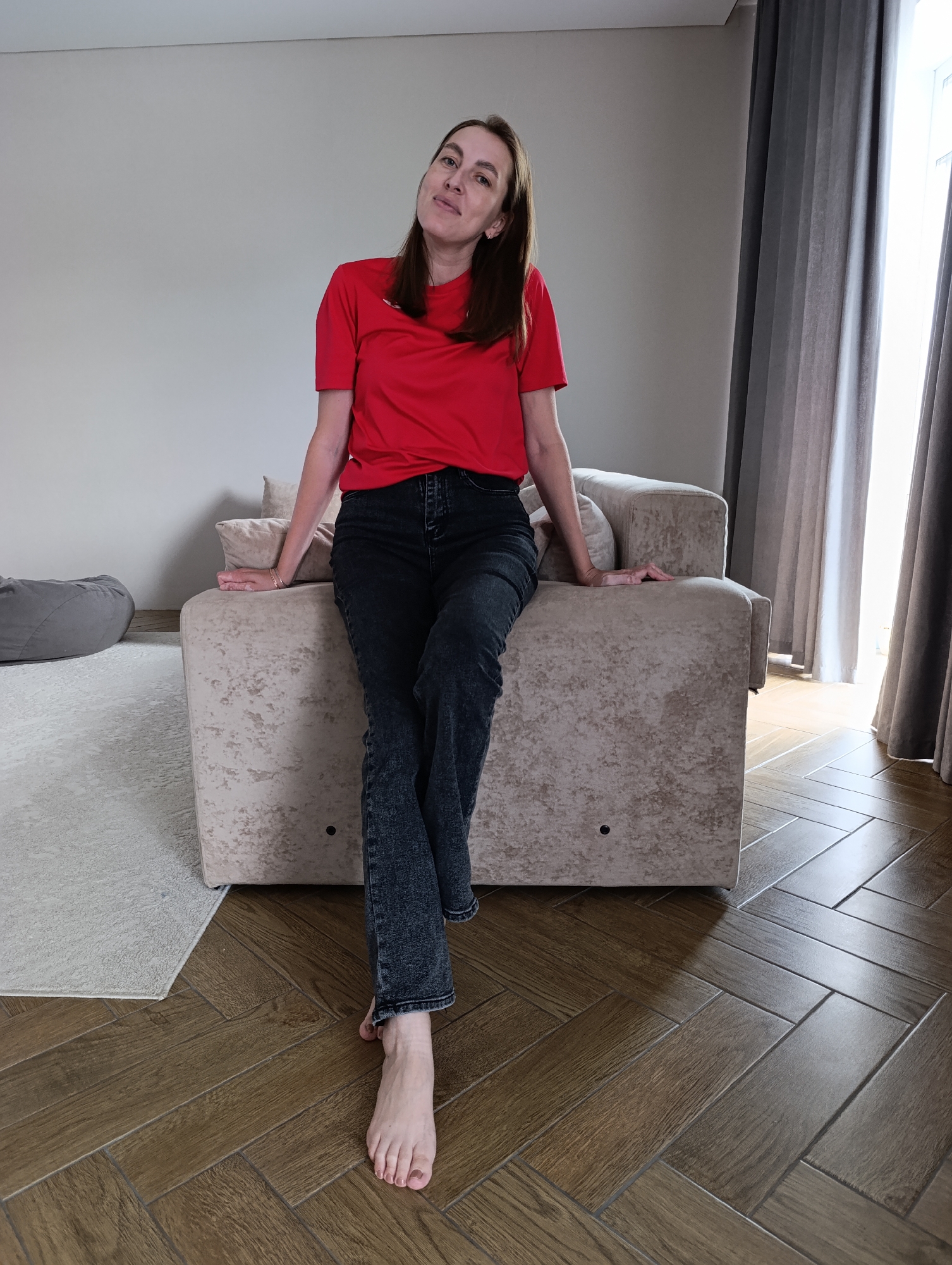 A woman with medium-length brown hair is sitting on the armrest of a beige sofa in a living room. She is wearing a red t-shirt and dark wash jeans. Her right hand is resting on the back of the sofa, and her left arm is bent with her hand on her hip. Her legs are crossed, and her bare feet are visible. The sofa is positioned on a herringbone-patterned wooden floor. To the left of the sofa, a light-colored rug is visible, with a grey beanbag chair beyond it. In the background, a light-colored wall and grey curtains covering a window can be seen. The scene is well-lit, suggesting it's daytime. The location is identified as Chaikovsky, Russia.