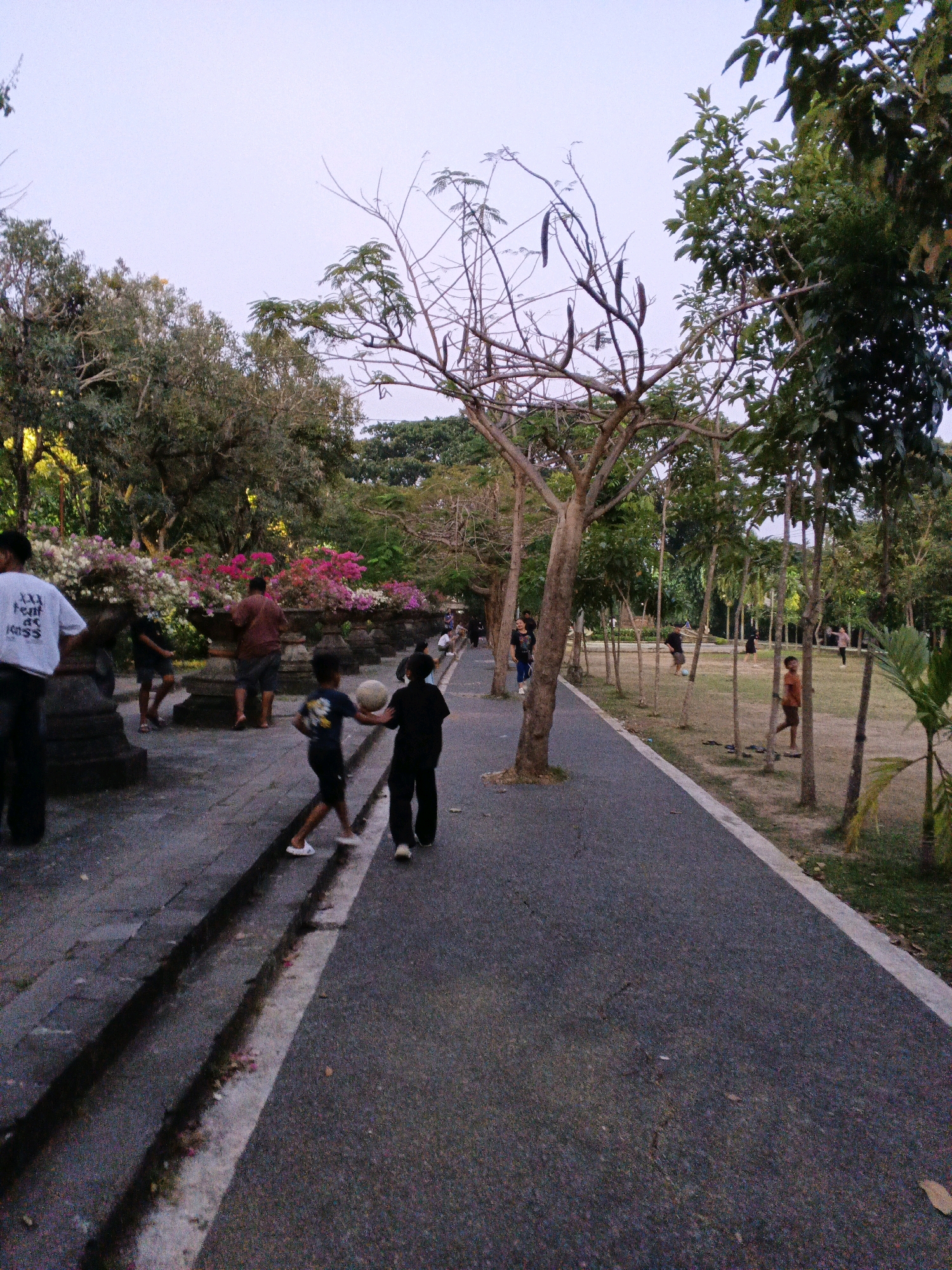 In Denpasar, Indonesia, at dusk, a paved pathway runs through a park lined with trees and foliage. Two young boys are playing with a white ball on the pathway. To the left of the pathway, a raised stone ledge is adorned with large planters filled with colorful flowers. Several adults are visible in the background, some walking and others standing near the planters. The sky above is pale, indicating the late hour of the day.