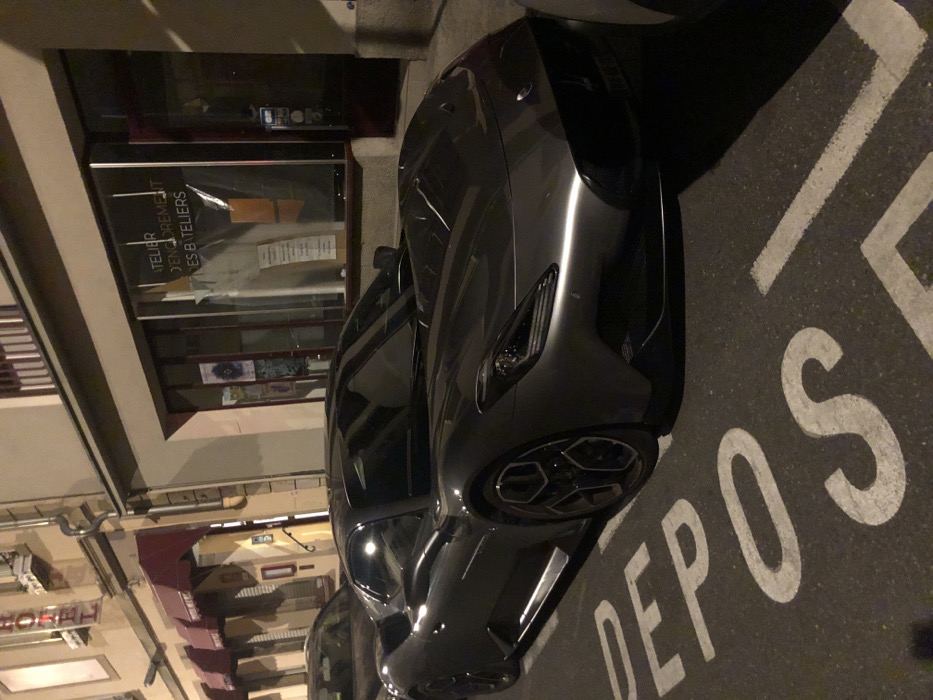 A dark metallic grey Maserati MC20 sports car is parked curbside on an asphalt street at night in Strasbourg, France. The vehicle's front and driver's side are prominently featured, positioned over white painted road markings that spell "DEPO" from a "DÉPÔT" (deposit/drop-off) zone. To the immediate left, an older building displays a vertical "HOTEL" sign and multiple red awnings. Directly behind the vehicle, a storefront with large glass windows shows interior items and text on the glass reading "ATELIER D'ENCOREMENT DES BATELIERS," indicating a framing workshop. The scene is illuminated by artificial streetlights, casting reflections on the car's surface. This location is identified as approximately 13 Rue des Bateliers, 67000 Strasbourg, France. No individuals are visible.
