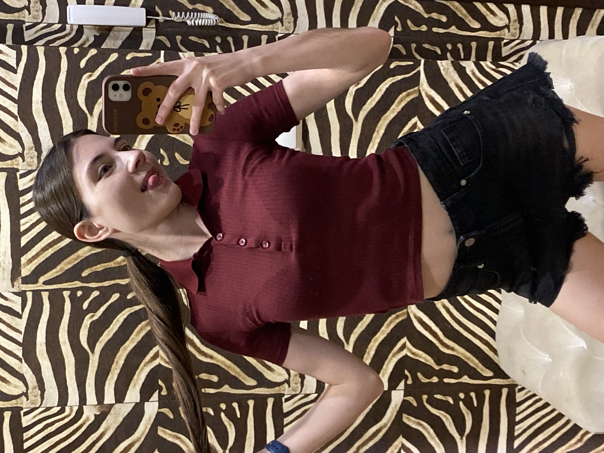 A woman takes a selfie while lying on a bed with a zebra-striped pattern. She wears a maroon, short-sleeved, collared shirt and black denim shorts. She is holding a brown phone with a teddy bear case. Her long hair is pulled back in a ponytail. A white telephone is visible in the upper left corner of the image. The location is not identifiable as Votkinsk, Russia, as there is no geographically specific information in the image.