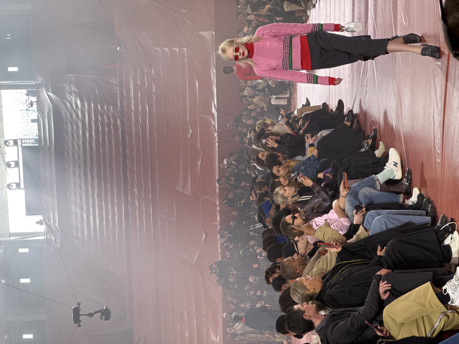 This image depicts a fashion show taking place inside the Accor Arena (Palais Omnisports de Paris-Bercy) in Paris, France. The arena's tiered seating is entirely draped in a reddish-orange fabric, creating both the runway and audience areas. In the foreground, a blonde female model walks on the reddish-orange runway, moving from the background towards the viewer's right. She wears a pink cable-knit cropped sweater with green and red stripes over a red turtleneck, dark grey cropped trousers, black loafers, and red rectangular sunglasses, while carrying a small white handbag in her left hand.
To her left, a dense crowd of spectators sits in multiple rows along the runway, facing the model. Many individuals are holding smartphones, actively photographing or video recording the event. Further back, more people are visible, including photographers equipped with professional cameras and tripods. Overhead, a cable-mounted camera system is suspended. The upper background reveals the arena's covered tiers extending upwards, with bright, diffused light sources visible at the top and a "DRAPEL" sign faintly discernible. The overall atmosphere appears slightly hazy, possibly due to stage lighting effects.