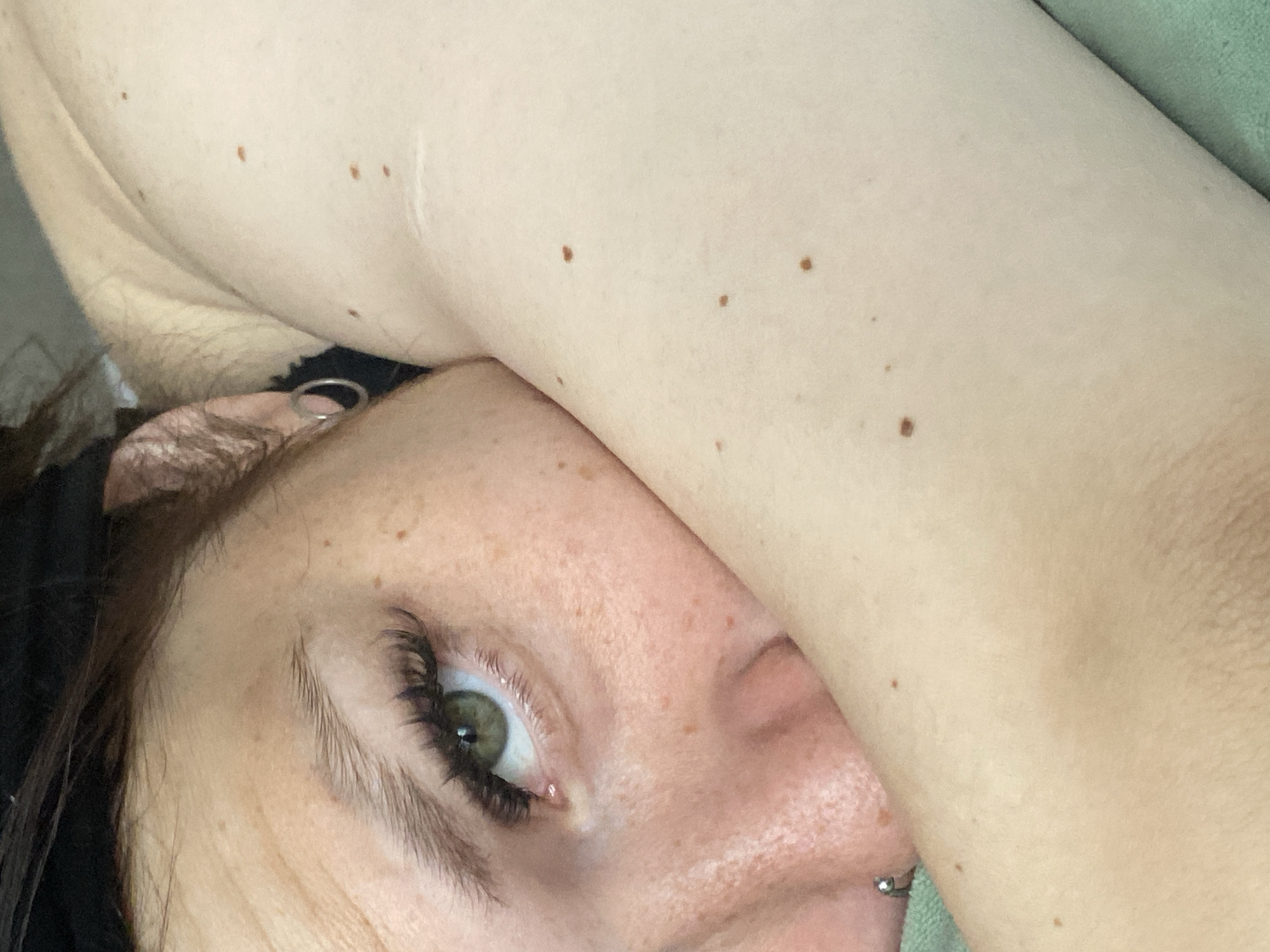 This is a close-up, high-angle shot of a person's face and arm. The individual has green eyes and is wearing false eyelashes. Their skin is fair with freckles. They are wearing a silver hoop earring in their left ear and a silver piercing on their nose. The person's arm is bent, covering part of their face. The background is blurred and appears to be a green fabric. The scene is not specific to any city and does not contain enough information to determine the location in Chaikovsky, Russia.