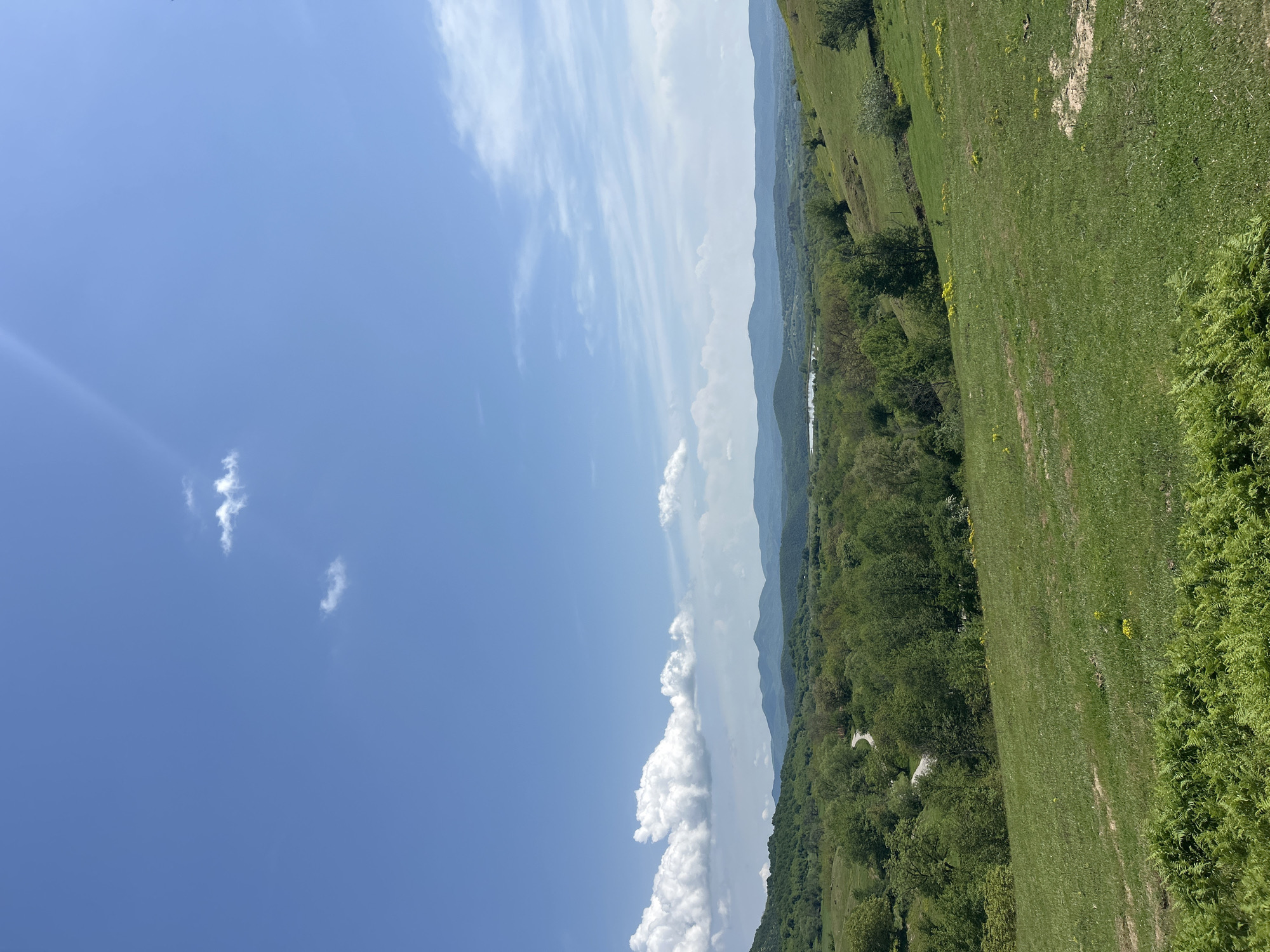 The image depicts a landscape in the city of Vasil Levski, Bulgaria, featuring a blue sky with scattered white clouds covering almost half of the sky. The terrain is green and slightly sloped, with a meadow and some forestation. The mountains can be seen in the distance.