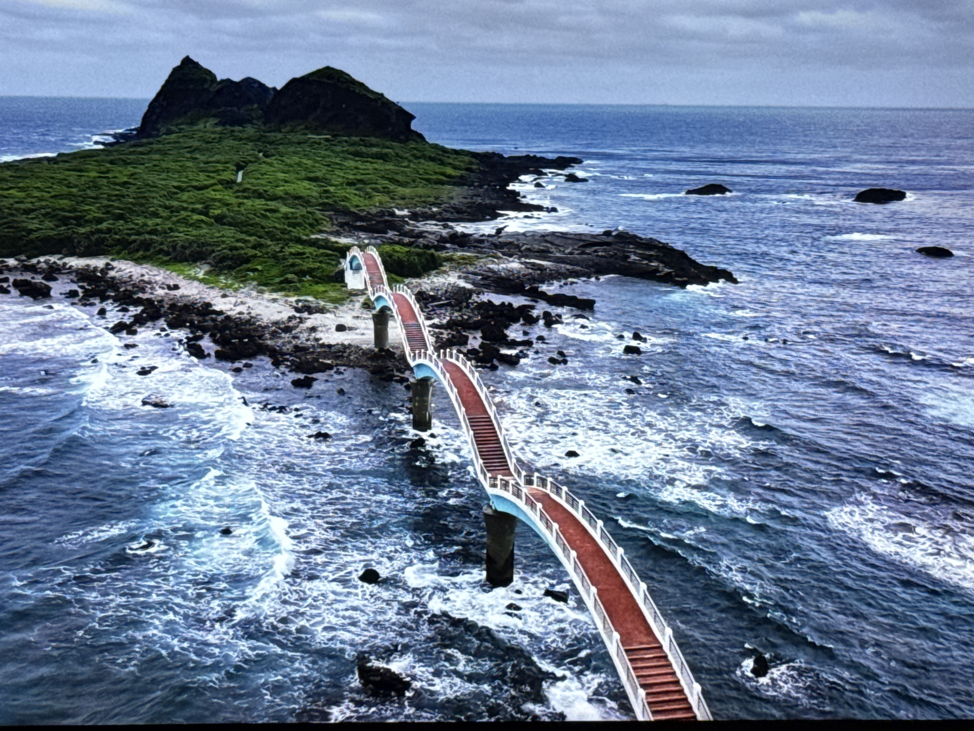 An aerial view captures a scenic bridge, painted in a vibrant red and white with blue accents, stretching over choppy, dark blue water. The bridge connects to a rocky, black-sand shoreline and leads towards a lush, green island with jagged peaks. The sky is overcast and a pale gray. The water is a deep blue, with white foam indicating waves crashing against the rocks and the bridge's supports. The island is densely covered in green vegetation, with a clear path visible winding up one of its slopes. The bridge appears to be a pedestrian walkway with railings and steps. This scene does not depict the city of Burgas, Bulgaria.