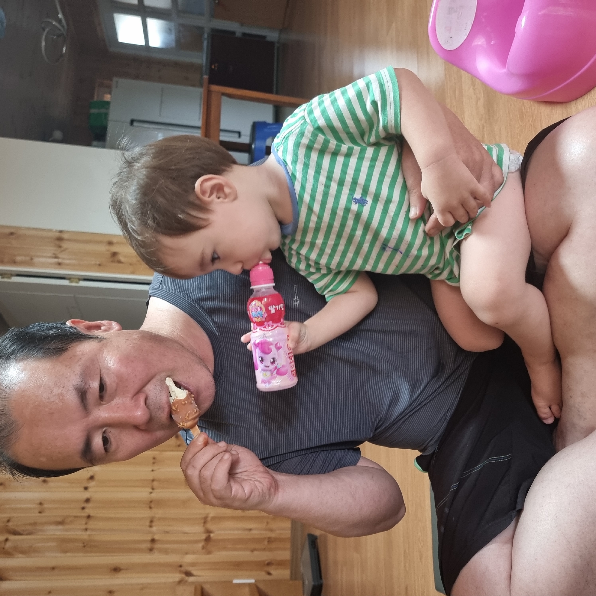 The image depicts an indoor scene likely within a residential setting. An adult male, seated on the floor, is eating a piece of food on a stick. He is holding a small pink bottle with a white cap and a pink label featuring a cartoon character, which a young child is drinking from. The child, dressed in a green and white striped shirt, is seated on the man's lap. A pink potty training seat is visible to the right of the child. The flooring appears to be wooden planks, and a wooden paneled wall is visible behind the man. Other objects in the background include what appear to be kitchen appliances and furniture, suggesting a living area or kitchen. The lighting suggests it is daytime. The overall impression is a casual, domestic moment.