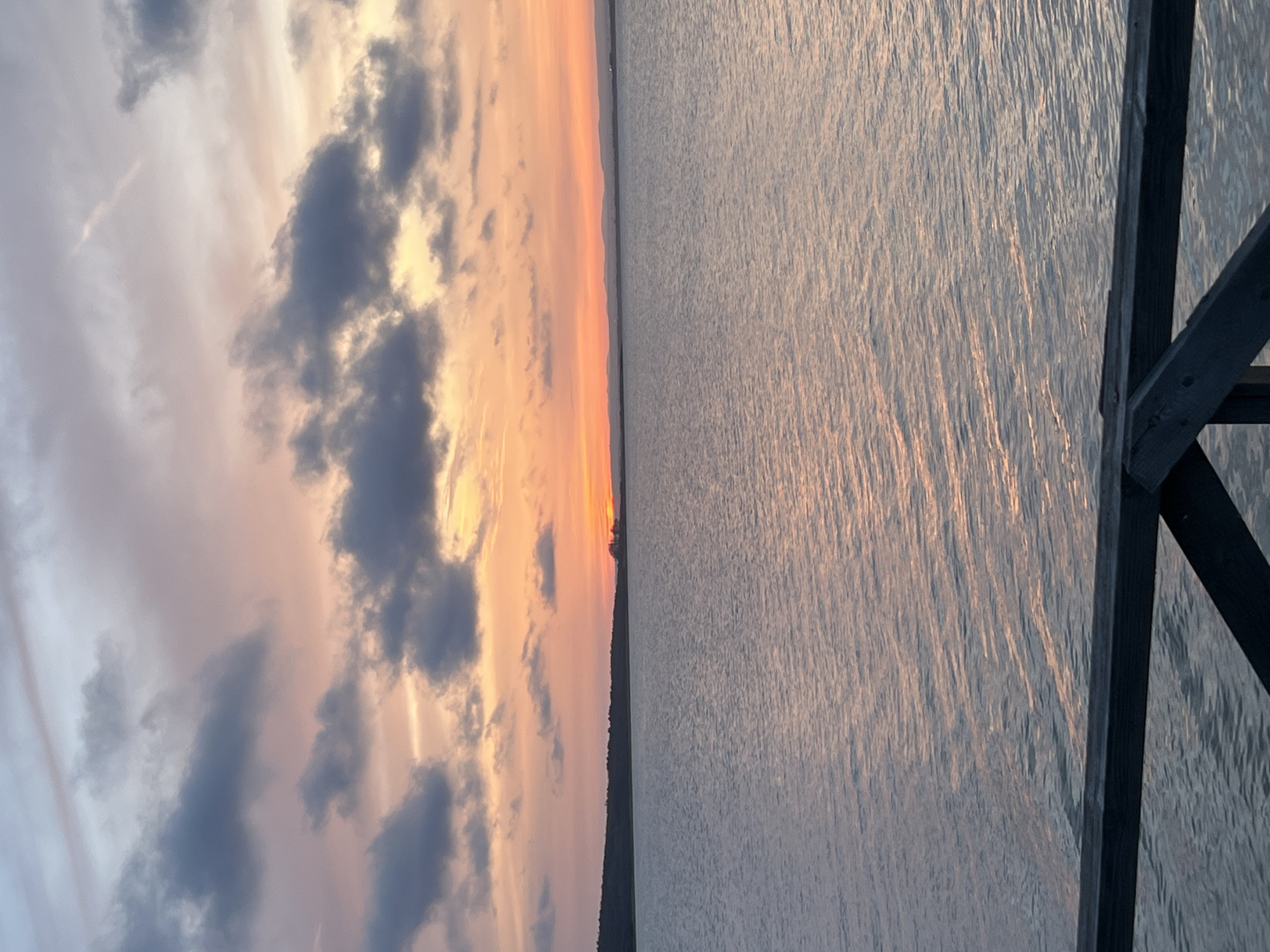 The image, rotated 90 degrees clockwise, captures a serene sunset or sunrise over a vast body of water, likely the Black Sea near Burgas, Bulgaria.
The left side of the frame is dominated by a dramatic sky, painted with soft pastel pinks, oranges, and yellows from the low-lying sun. Scattered across the sky are numerous clouds; some appear as dark, shadowed forms, while others are brightly illuminated by the warm light, blending into the colorful canvas. The sun itself is partially visible, a bright orange orb just peeking above the distant, dark silhouette of a land or treeline that forms the horizon, stretching across the middle of the image.
The right half of the image features the expansive body of water, its surface textured with countless small ripples and waves. These water movements catch and reflect the warm light from the sky, creating shimmering streaks of orange and pink against the cooler blue-gray tones of the water. In the far right foreground, a dark, angular wooden structure, possibly part of a pier or dock, protrudes into the scene, its sturdy, shadowed beams providing a contrasting element to the soft, ethereal colors.
The overall scene conveys a sense of calm and natural beauty. There are no people visible, and no text is present in the image.