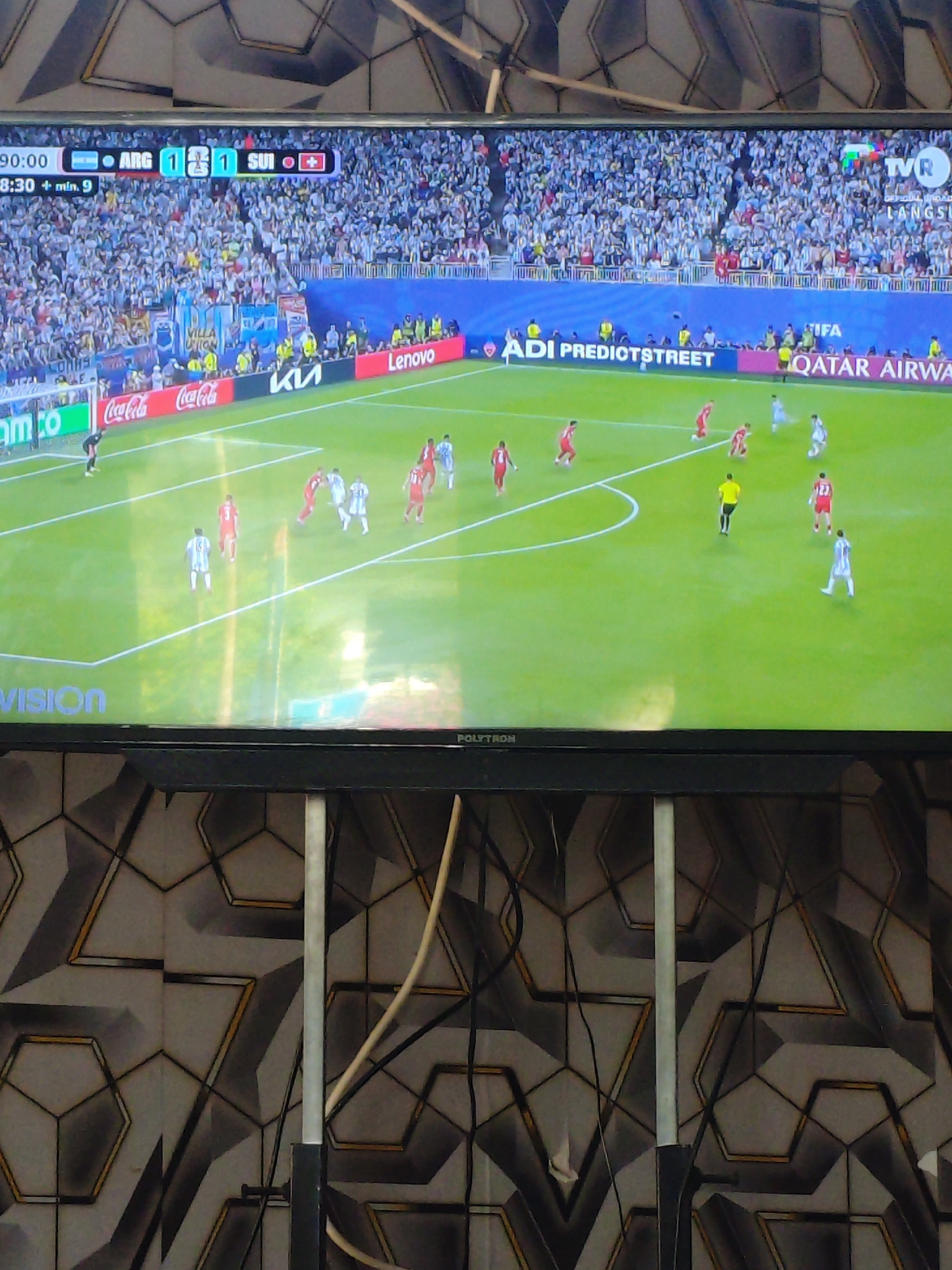 The image shows a television screen displaying a soccer match. The score displayed is 1-1 between ARG and SUI. The game time is 90:00 + min. 9. The broadcast is identified as TVR. There are advertisements along the side of the field for Coca-Cola, Kia, Lenovo, ADI PREDIOTSTREET, and QATAR AIRWAYS. Several players in white and red uniforms are visible on the green soccer field. The television set itself is a black flat screen with the brand name POLYTRON visible at the bottom. The background behind the television is a patterned wall with geometric designs in black, grey, and yellow. The overall environment suggests a living room or entertainment area in Parangsembilan, Indonesia, where this scene is being viewed.