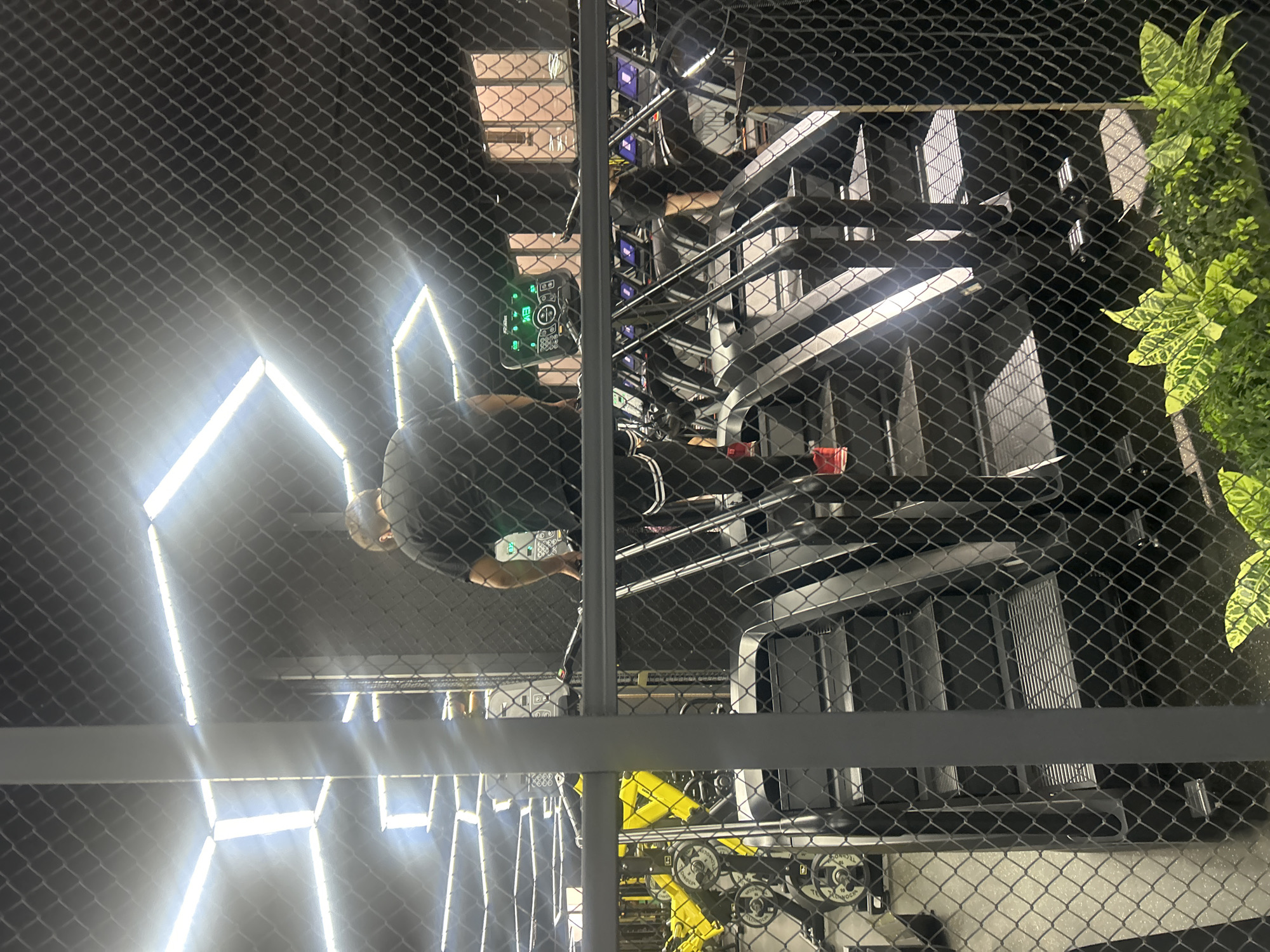 The image depicts a gym interior, likely in Almaty, Kazakhstan, viewed through a chain-link fence. A person, viewed from behind, is climbing a set of stairs on a stair climber machine. They are wearing a black t-shirt and black athletic pants with white stripes. The gym features modern, geometric lighting fixtures on the ceiling, casting bright light on the scene. In the background, other gym equipment, including weight machines and treadmills with illuminated screens, are visible. The overall environment appears to be a contemporary fitness facility.