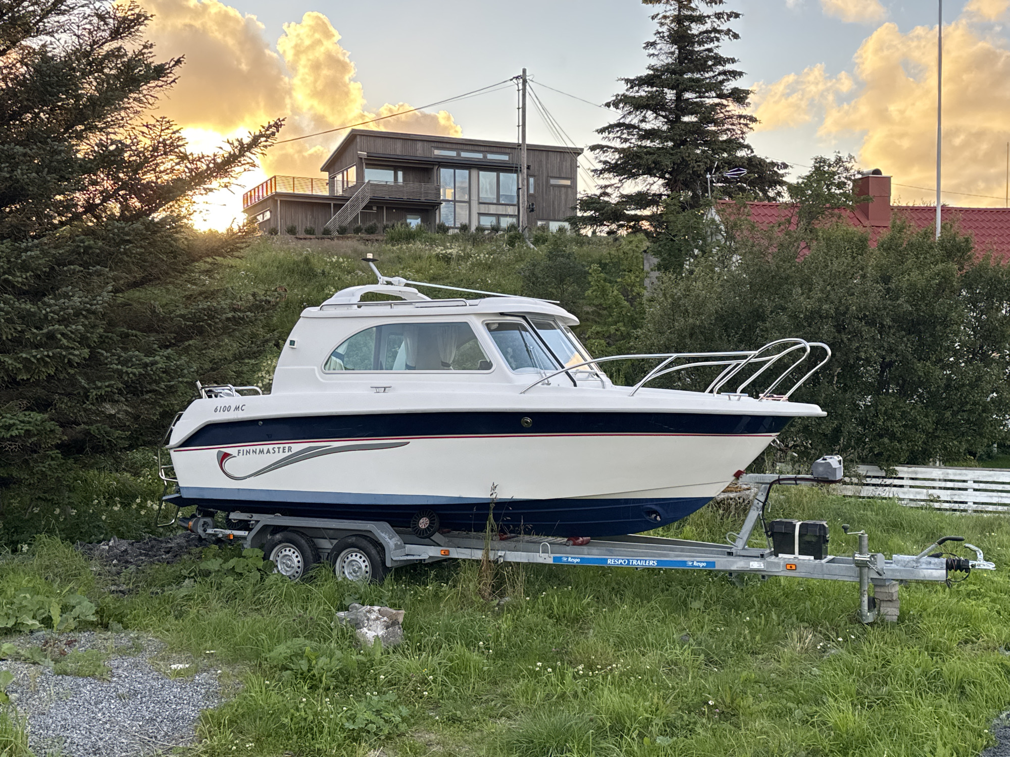 The image shows a white and blue Finnmaster boat on a trailer. The boat is parked on a grassy area with some gravel and rocks. In the background, there is a modern wooden house with large windows, situated on a hillside. A tall evergreen tree stands to the right of the house, and another tree is visible to the left. The sky is filled with dramatic clouds illuminated by the setting sun, creating a warm, golden light. The scene appears to be in a natural, possibly rural or suburban, setting. The boat has the model number "6100 MC" visible on its stern. The trailer is labeled "RESPO TRAILERS". The image captures a peaceful moment, likely in the evening, given the lighting conditions. The specific location is Lovund, Norway, as indicated by the context.
