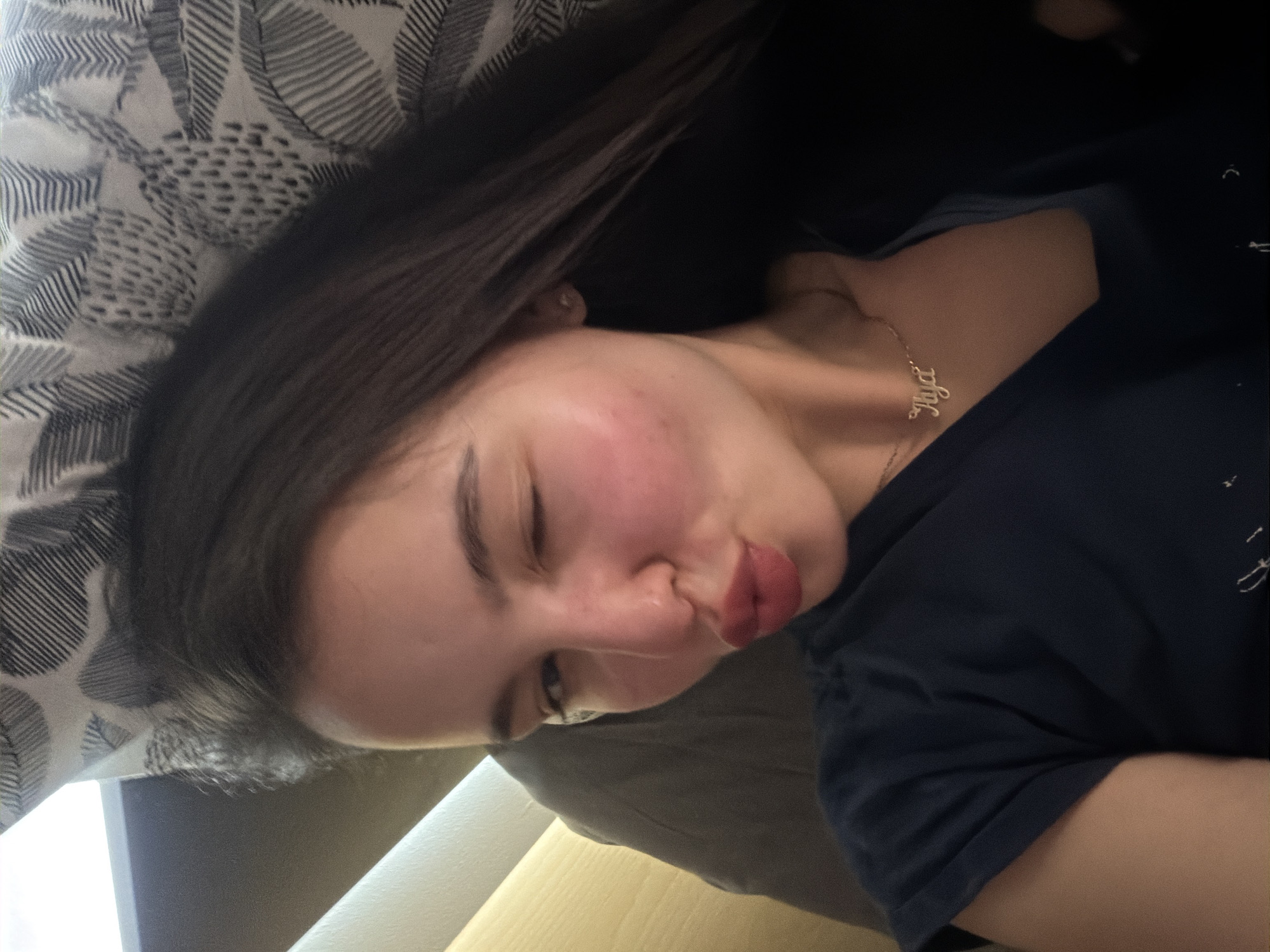 A young woman is lying on a patterned pillow. She is wearing a dark-colored t-shirt and a necklace with a name pendant. Her eyes are closed, and her lips are pursed. The background includes a wooden surface. The location is Unknown, Unknown.