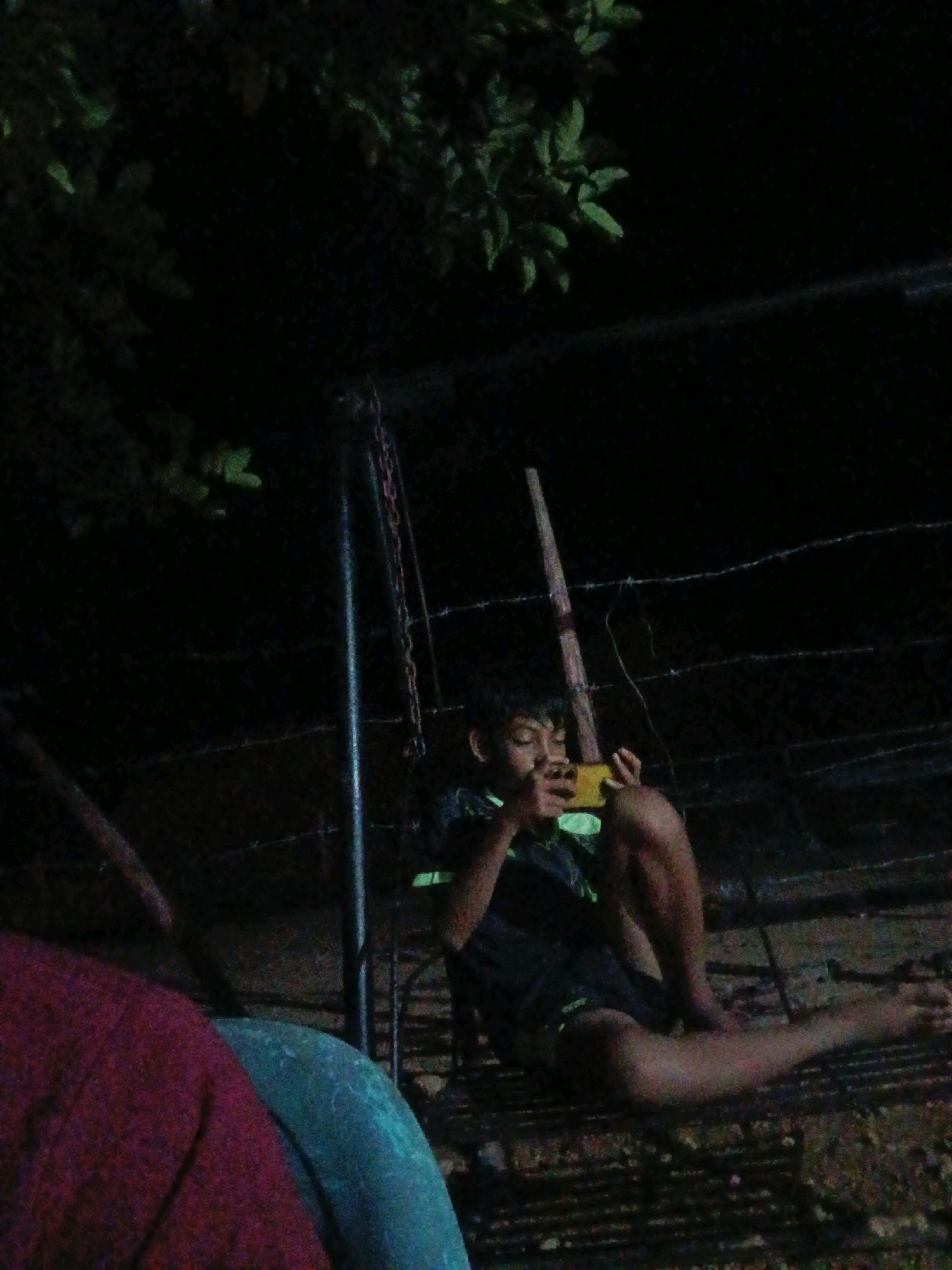 A young person is sitting on a metal structure outdoors at night, looking at a yellow smartphone. The person is wearing a dark t-shirt with some green lettering and dark shorts. They are sitting with their legs extended. Behind them, a chain hangs from what appears to be a swing set frame, and barbed wire is strung across the background. Some leafy branches are visible in the upper left corner, illuminated by an unseen light source. The ground beneath the person appears to be dirt and possibly some scattered debris. The overall lighting suggests it is nighttime, with the only significant light coming from the smartphone screen. The location is Brangan Mulya, Indonesia.