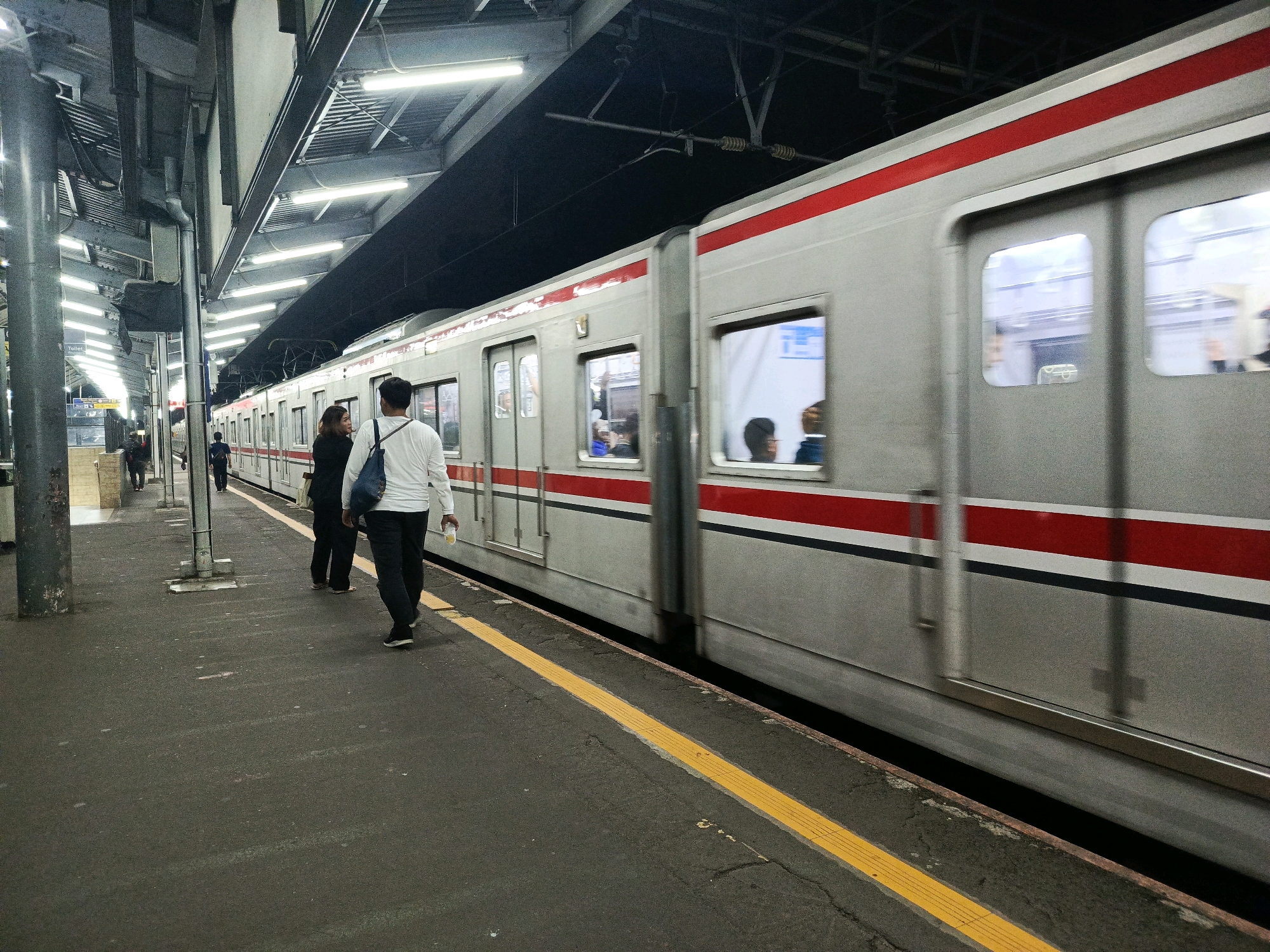 A commuter train, identified by its silver and red stripe livery, is captured in motion at a dimly lit platform in South Jakarta, Indonesia. The train is partially visible, its windows reflecting the station's fluorescent lighting. On the platform, several individuals are present. In the foreground, two people are walking away from the camera; one, wearing a white long-sleeved shirt and dark trousers, carries a blue bag over their shoulder and a light-colored cup in their right hand. The other person, in dark clothing, walks slightly ahead. Further down the platform, another individual in dark clothing is walking towards the left of the frame, and beyond them, more figures are indistinctly visible. The platform itself is asphalt, marked with a prominent yellow tactile paving strip along the edge. Overhead, a complex structure of beams and conduits supporting the station's roof and lighting system is evident. The overall atmosphere is that of a typical urban railway station during evening hours.