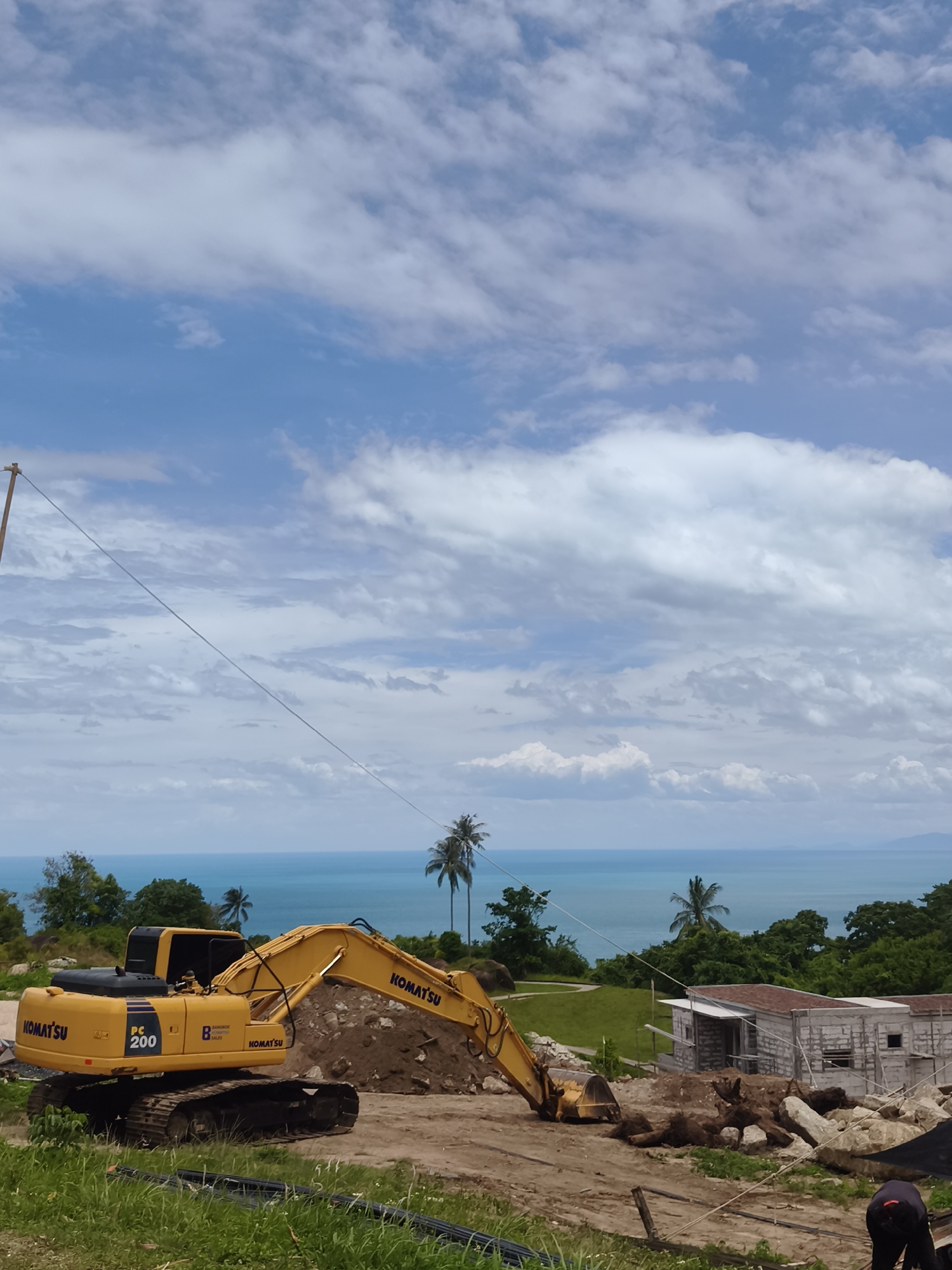 A yellow Komatsu PC 200 excavator is positioned on a dirt construction site overlooking the ocean in the Ko Samui District of Thailand. The excavator is actively engaged in moving a large pile of dirt and debris. In the background, the turquoise ocean stretches to the horizon, dotted with islands and framed by lush green trees and palm trees. A partially constructed building with concrete block walls is visible to the right of the excavator. A person wearing dark clothing is partially visible in the lower right corner of the frame, appearing to be working at the construction site. The sky is a mix of blue with scattered white clouds. The foreground consists of grassy areas and some black piping.