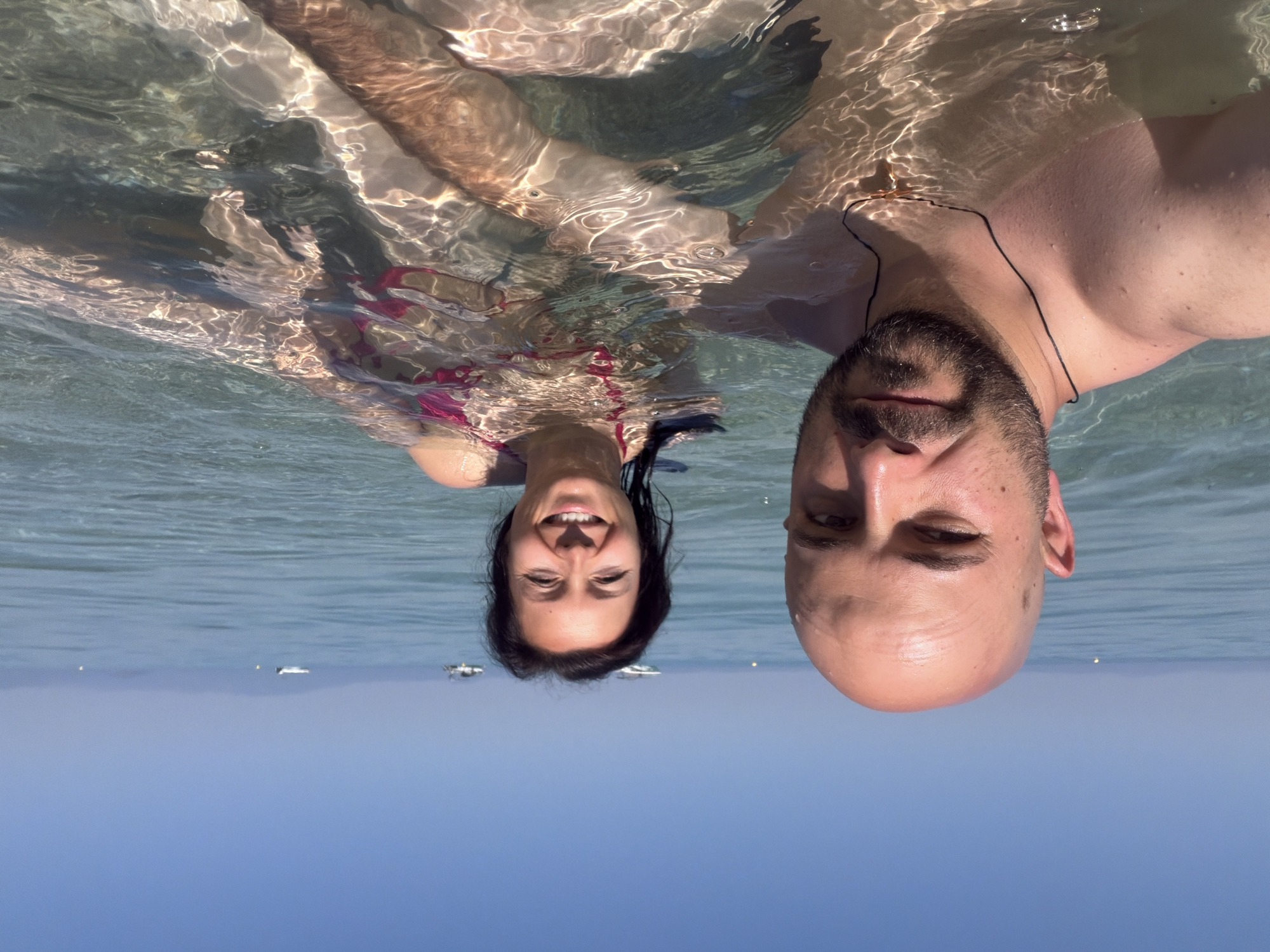 In the city of Pallini Municipal Unit, Greece, a man and a woman are captured in a close-up selfie while submerged in clear, shallow ocean water under a bright, cloudless sky. The man, with a shaved head and a beard, is positioned in the foreground, looking towards the camera with a slight smile. He is wearing a dark string necklace with a golden pendant. The woman, with dark hair and a beaming smile, is situated behind and to his right, also looking at the camera. She is wearing a red bikini top. The water's surface is disturbed by ripples, creating a dappled light effect on their skin and the surrounding water. In the background, the calm sea stretches towards a distant, hazy coastline, with a few small boats visible on the horizon. The overall scene conveys a relaxed and joyful moment in a seaside environment.