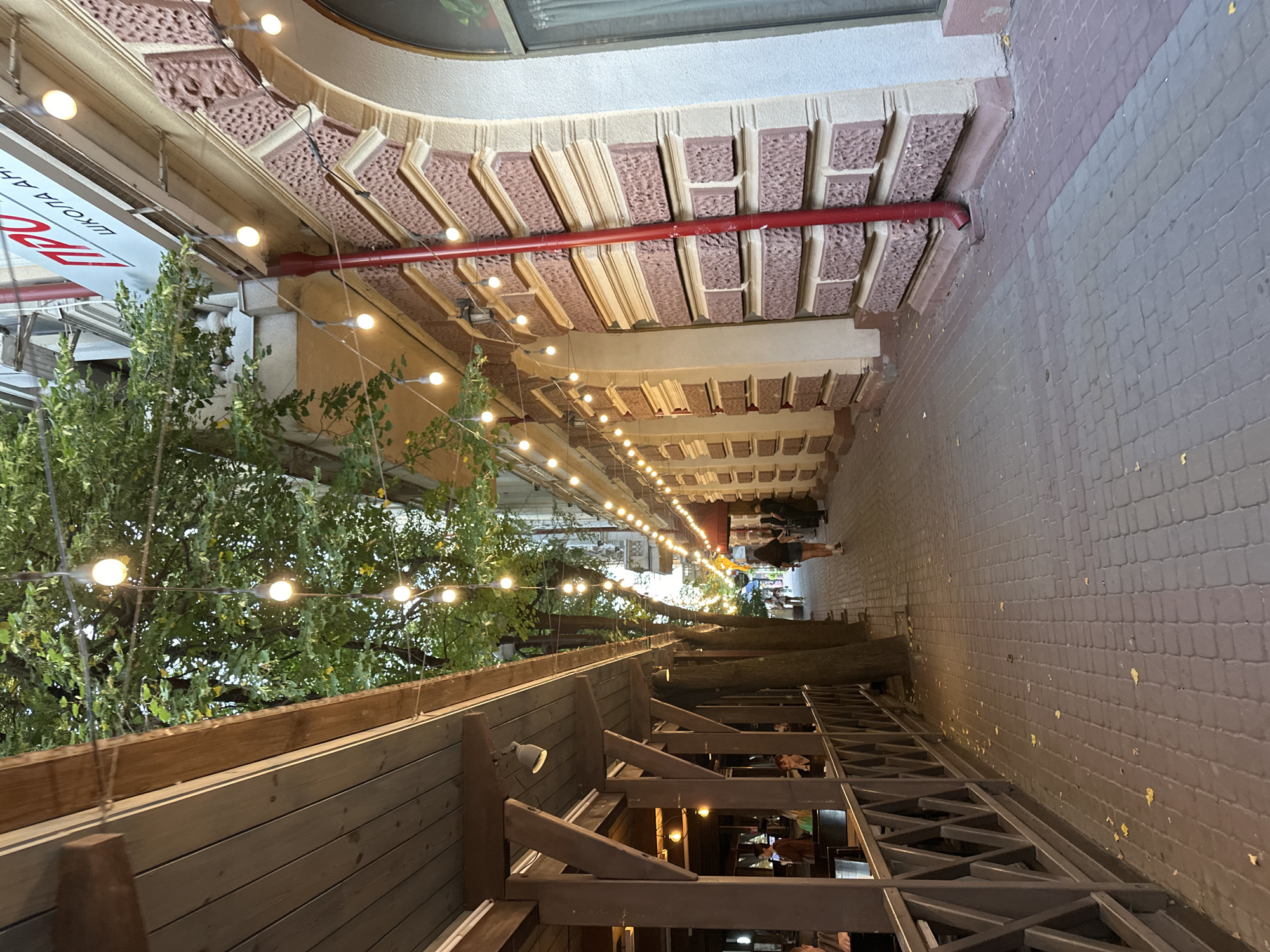 The image depicts a street scene in Ternopil, Ukraine. A pedestrian walkway paved with brick pavers runs along the right side of the image, bordered by a building with ornate architectural details. String lights with illuminated bulbs hang over the walkway, creating a festive atmosphere. Trees with green foliage are interspersed along the path. On the left side, there is an outdoor dining area with a wooden roof and railing. People are visible in the background, walking along the street and seated at tables in the dining area. The building on the right has a red downspout and a sign with Cyrillic lettering. The overall impression is a charming and lively urban setting, possibly a street lined with cafes and shops.