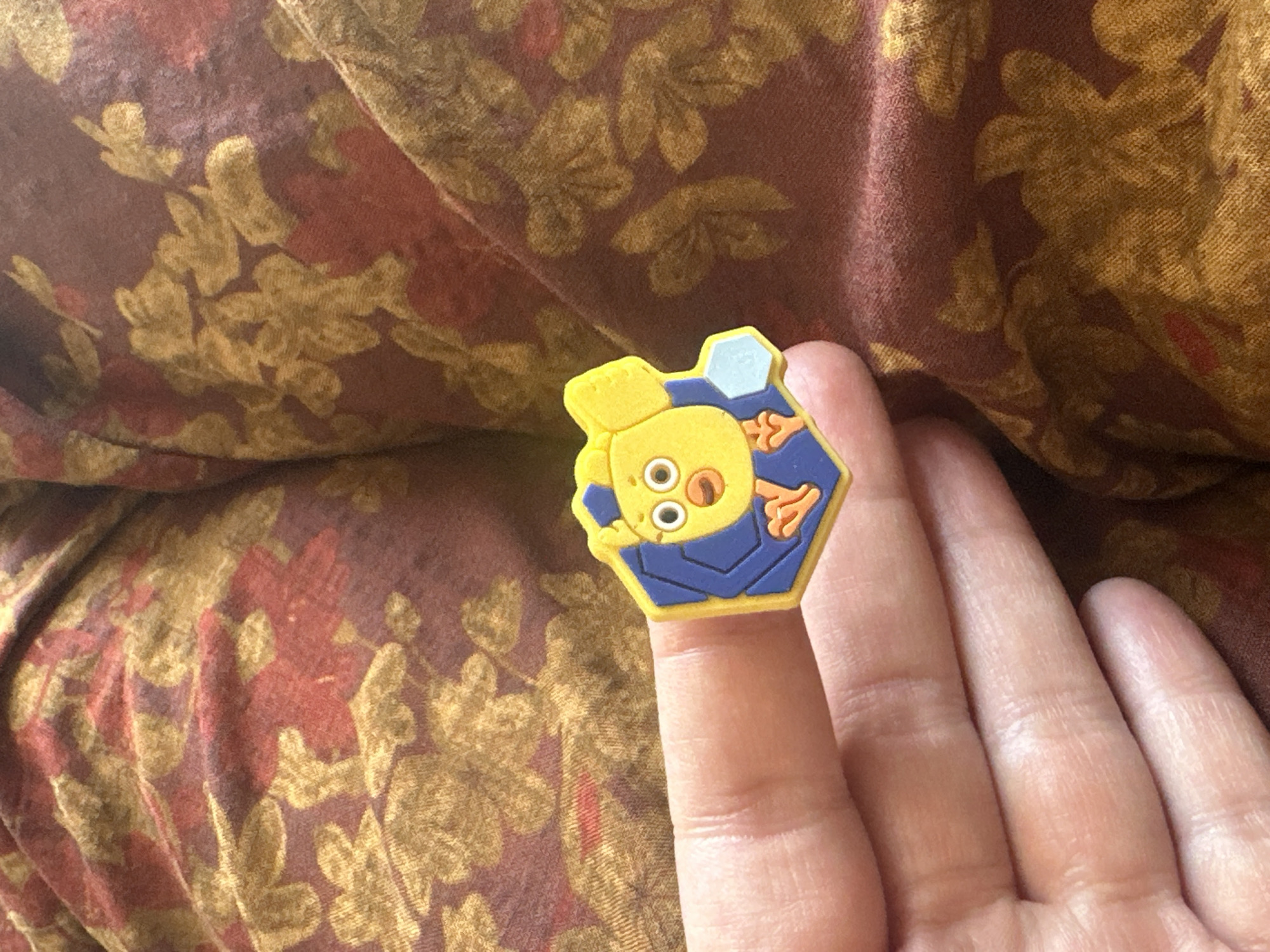 A close-up, eye-level shot shows a person's index finger holding a small, hexagonal charm with a cartoon chicken design. The charm is yellow with a blue background and orange accents. The person's finger is partially visible and appears to be light-skinned. The charm is positioned in the lower center of the frame. The background consists of a textured, patterned fabric in shades of burgundy and gold, with floral motifs. The scene appears to be indoors, with soft lighting. The location is stated to be in the village of Golovatsky, Kazakhstan, though this information cannot be visually verified from the image itself.