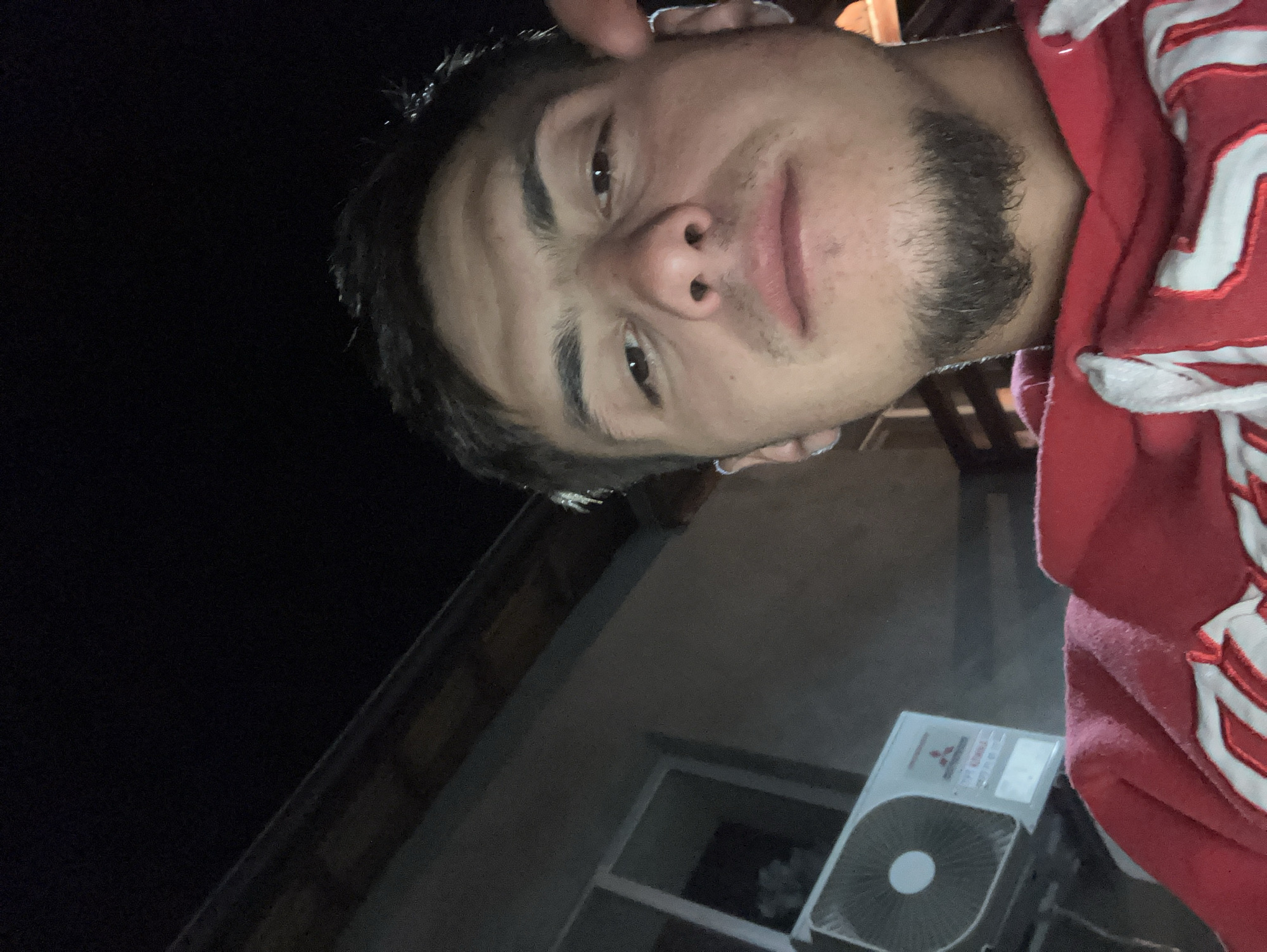 A close-up, dimly lit selfie shows a young man with brown hair and eyes, light skin, and a beard, wearing a red and white hooded sweatshirt. He appears to be standing outside at night. Behind him is a building with an air conditioning unit visible. There is no verifiable way to determine the location of this picture, nor can it be ascertained that the picture was taken in Komuniga, Bulgaria. The lighting is poor.