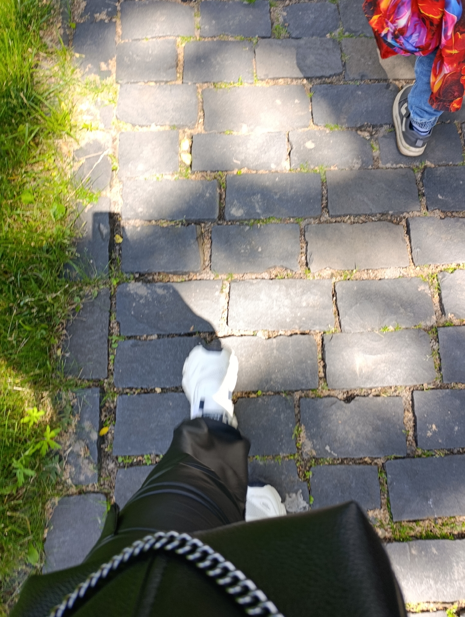 The image shows a view from above of a person walking on a cobblestone path. The cobblestones are dark gray and rectangular, with some grass growing between them. To the left of the path is a patch of bright green grass with some small plants. The person is wearing white sneakers, black pants, and carrying a black handbag with a silver chain strap. Another person, partially visible at the top right of the image, is wearing blue jeans and a colorful jacket with red, blue, and yellow patterns. The lighting suggests it is daytime, with some shadows cast on the path, indicating sunlight. The scene is set outdoors on a paved walkway, likely in a park or public area, possibly in Kyiv, Ukraine, given the context.