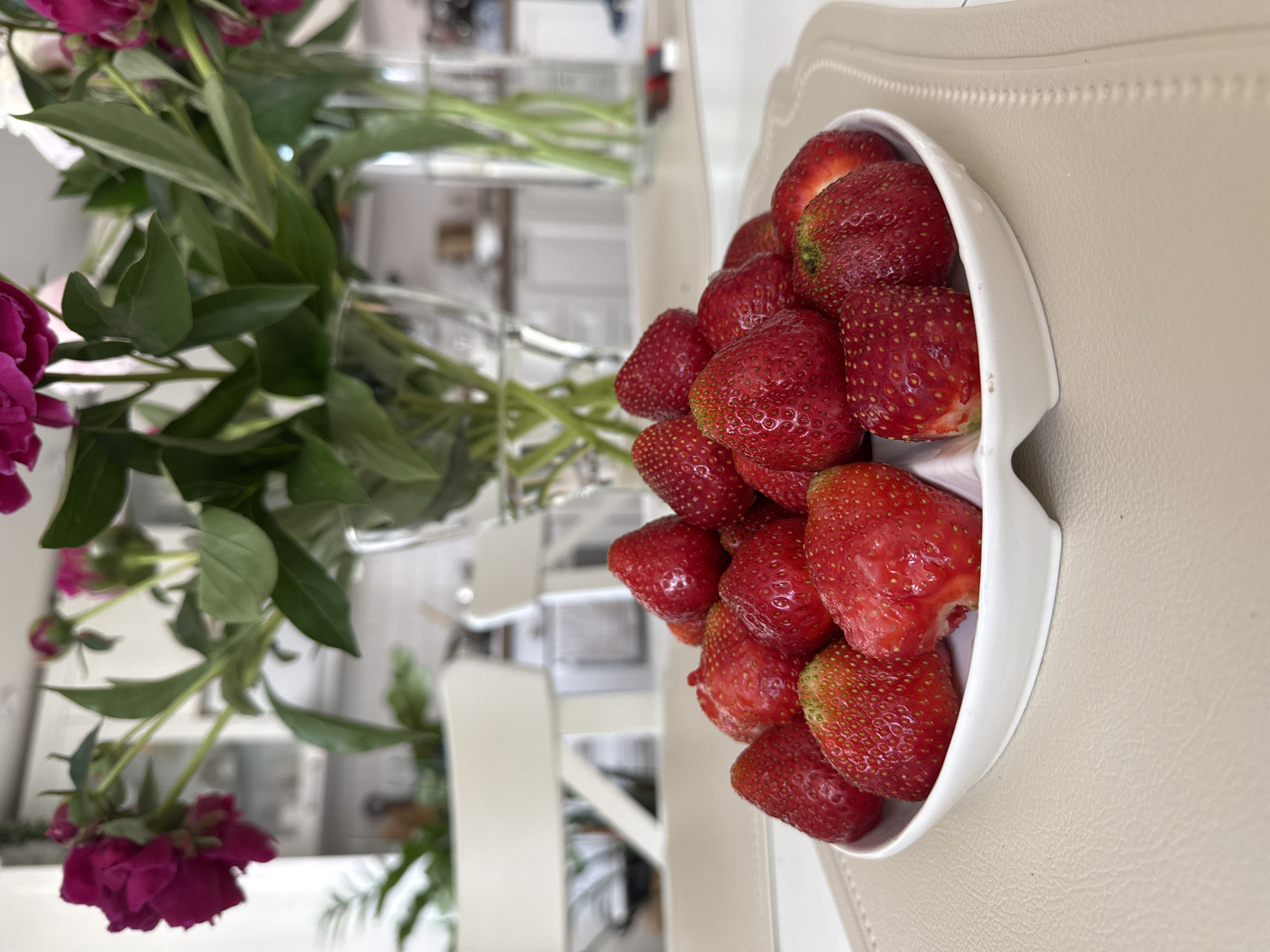 A close-up, eye-level, medium shot captures a white, divided serving dish filled with fresh strawberries, placed on a light beige placemat. The strawberries are vibrant red, some with green stems and leaves still attached. In the blurred background, a bouquet of dark pink peonies in glass vases rests on a white surface, with hints of kitchen cabinetry and chairs. The scene appears to be an indoor setting, likely a dining area or kitchen. There is no indication of the city of Chaikovsky, Russia, within the image or its provided crops.