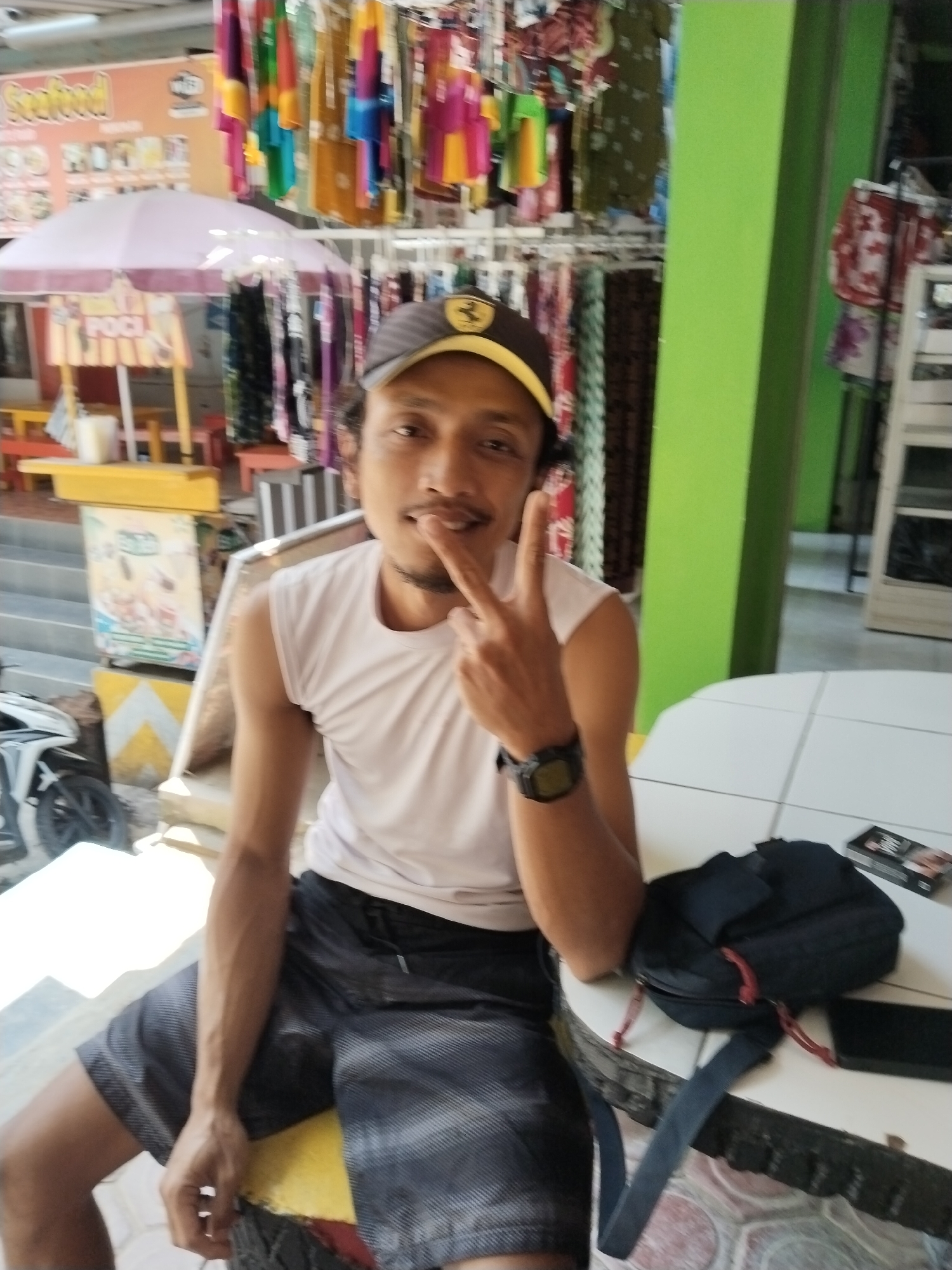 A man with a mustache and a beard, wearing a white sleeveless shirt, dark patterned shorts, and a brown baseball cap with a yellow brim, is sitting at a white tiled table outdoors. He is making a peace sign with his right hand. Behind him, a row of colorful clothes hangs on display racks, and a green pillar is visible. To his left, a pink umbrella is set up over a yellow stall with signage that includes the word "Seafood." A white scooter is parked to the left of the stall. A black backpack rests on the table in front of the man. The scene appears to be in a market or shop area in Prigi, Indonesia.