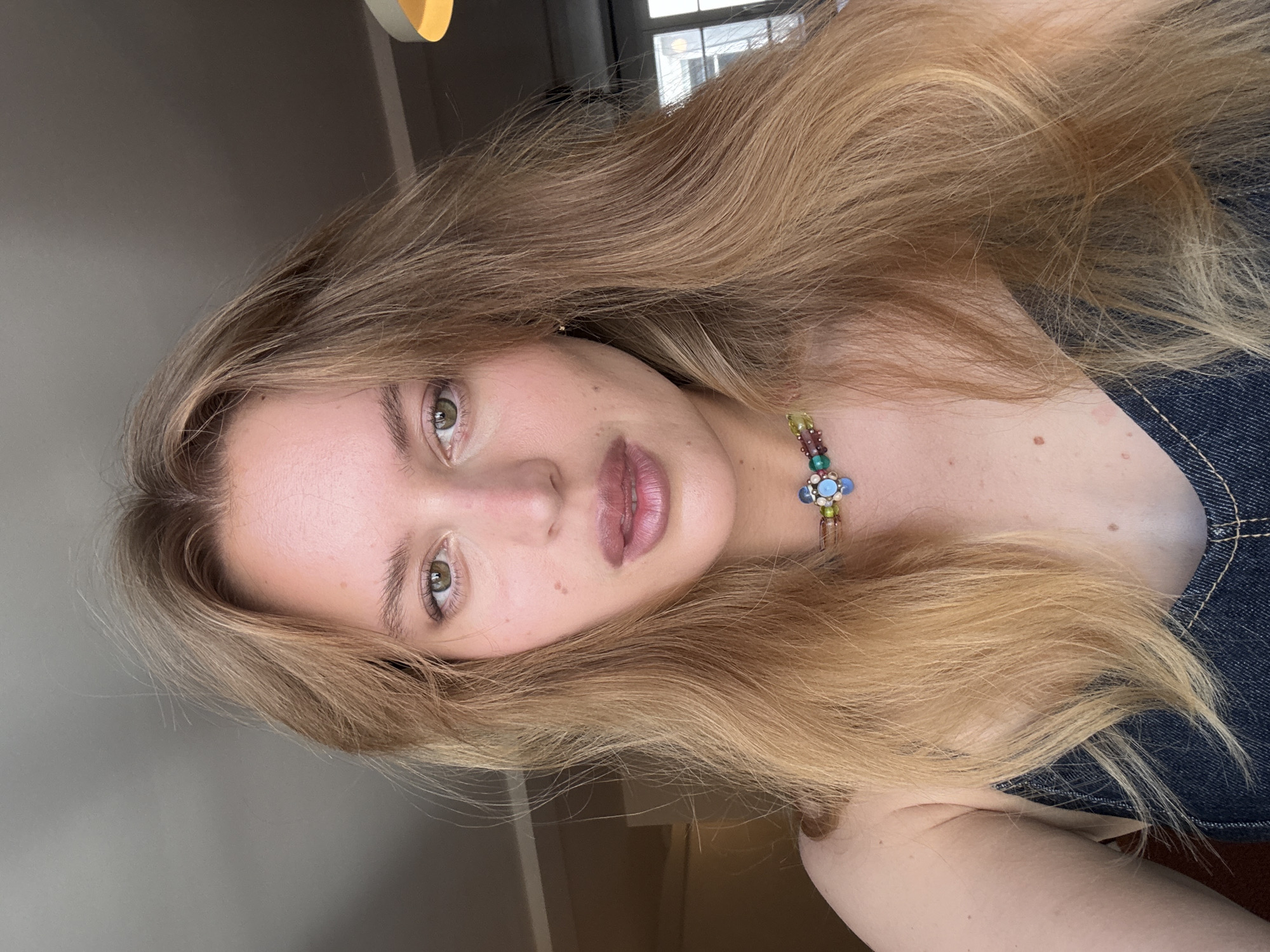 A close-up portrait, originally oriented on its side, features a young woman with long, wavy, light blonde hair cascading around her face and shoulders. Her gaze is directed calmly towards the camera, revealing green or hazel eyes. Her lips are a natural light pink hue. She has clear, fair skin, with a few subtle freckles visible.
Around her neck, she wears a distinctive, colorful beaded necklace, featuring various small beads in shades of green, purple, and yellow, culminating in a central, intricate charm with blue and green components, possibly resembling a flower. She is dressed in a dark blue garment, likely denim, visible on her right shoulder and chest.
The background is softly blurred, indicating an indoor setting. To her left, a plain, light grey wall is visible, while to her right, a window allows a glimpse of diffused daylight and reflections of an urban environment, suggesting buildings outside. A light-colored fixture is partially visible in the upper background. The natural lighting suggests the photo was taken during the daytime. The woman's expression is neutral and composed. The photograph was taken in Paris, France. No visible text is present in the image.