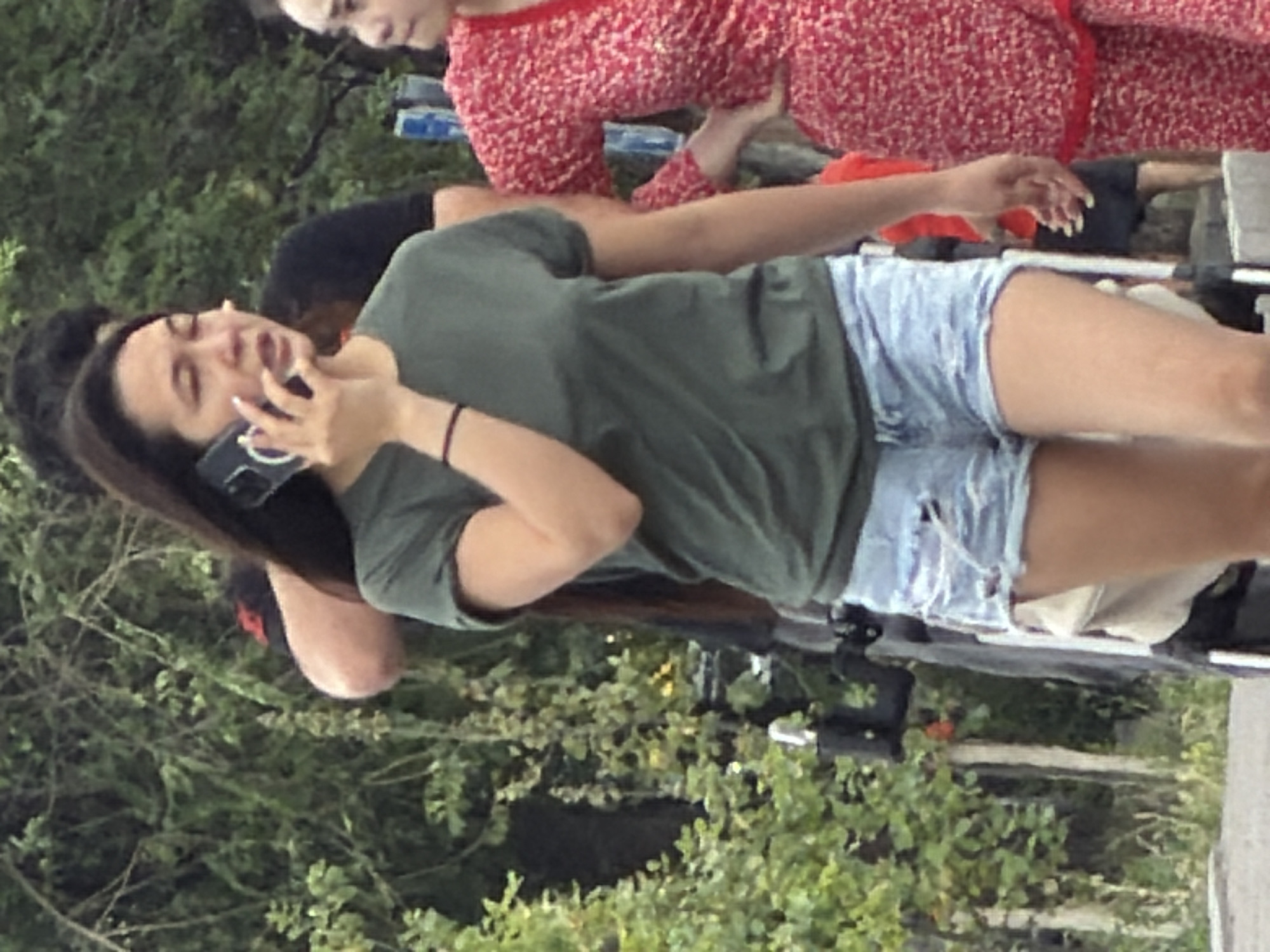 In an outdoor setting with trees in the background, a young woman wearing a green t-shirt and denim shorts is holding a cell phone to her ear and speaking. She has dark hair and appears to be in motion. To her right, another woman in a red floral dress is partially visible. Other individuals and a stroller are also present in the background, suggesting a public area such as a park or street. The lighting indicates it is daytime.