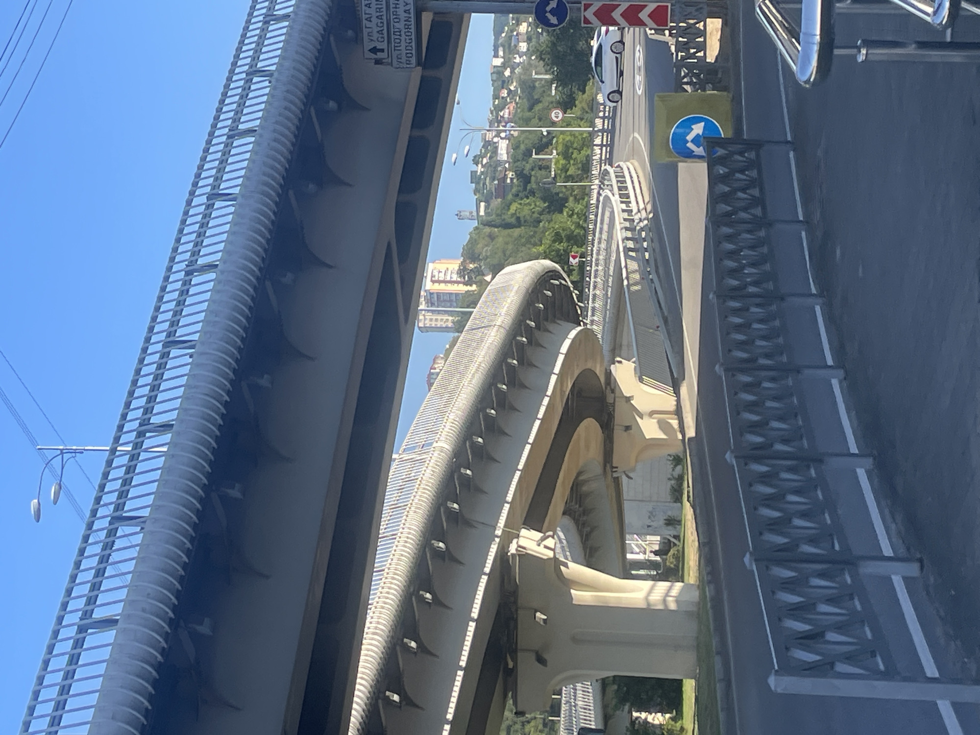 The image captures a multi-level overpass system in Sochi, Russia, under a clear blue sky. The foreground features a gray metal fence with an intricate, geometric pattern, partially obscuring a street with white lane markings. A large concrete overpass dominates the mid-ground, curving gracefully and supported by substantial pillars. Below this, another section of the overpass, seemingly older and more weathered, is visible.
To the right, a street curves around a grassy embankment. A white sedan is parked on the right side of the road, and a pedestrian crossing sign with blue and white arrows is affixed to a yellow cylindrical base. A red and white striped traffic delineator stands near the sign. Further down the road, a circular speed limit sign displaying "40" is mounted on a pole.
In the background, the city of Sochi unfolds on a hillside dotted with buildings of various sizes and architectural styles, some appearing modern and others more traditional. Lush green trees cover the slopes, indicating a temperate climate. A tall, slender tower, possibly a water tower or communication mast, is visible on the distant hilltop. The overall scene depicts a modern urban infrastructure integrated into a natural, hilly landscape.