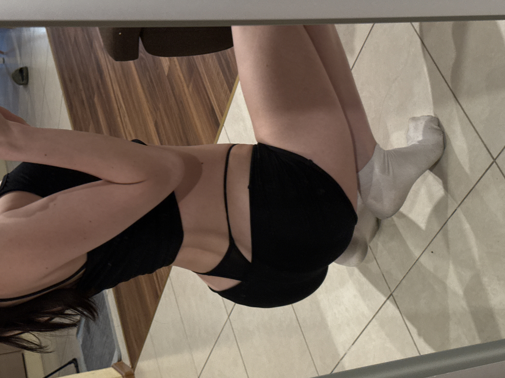 The image shows a person in a black sports bra and black shorts, wearing white socks. The person is standing on a tiled floor, with a wooden floor visible in the background. The setting appears to be indoors, possibly a home or gym. The lighting suggests it could be daytime. No specific activity or event is clearly depicted, but the attire suggests a casual or athletic context. There is no visible text in the image.