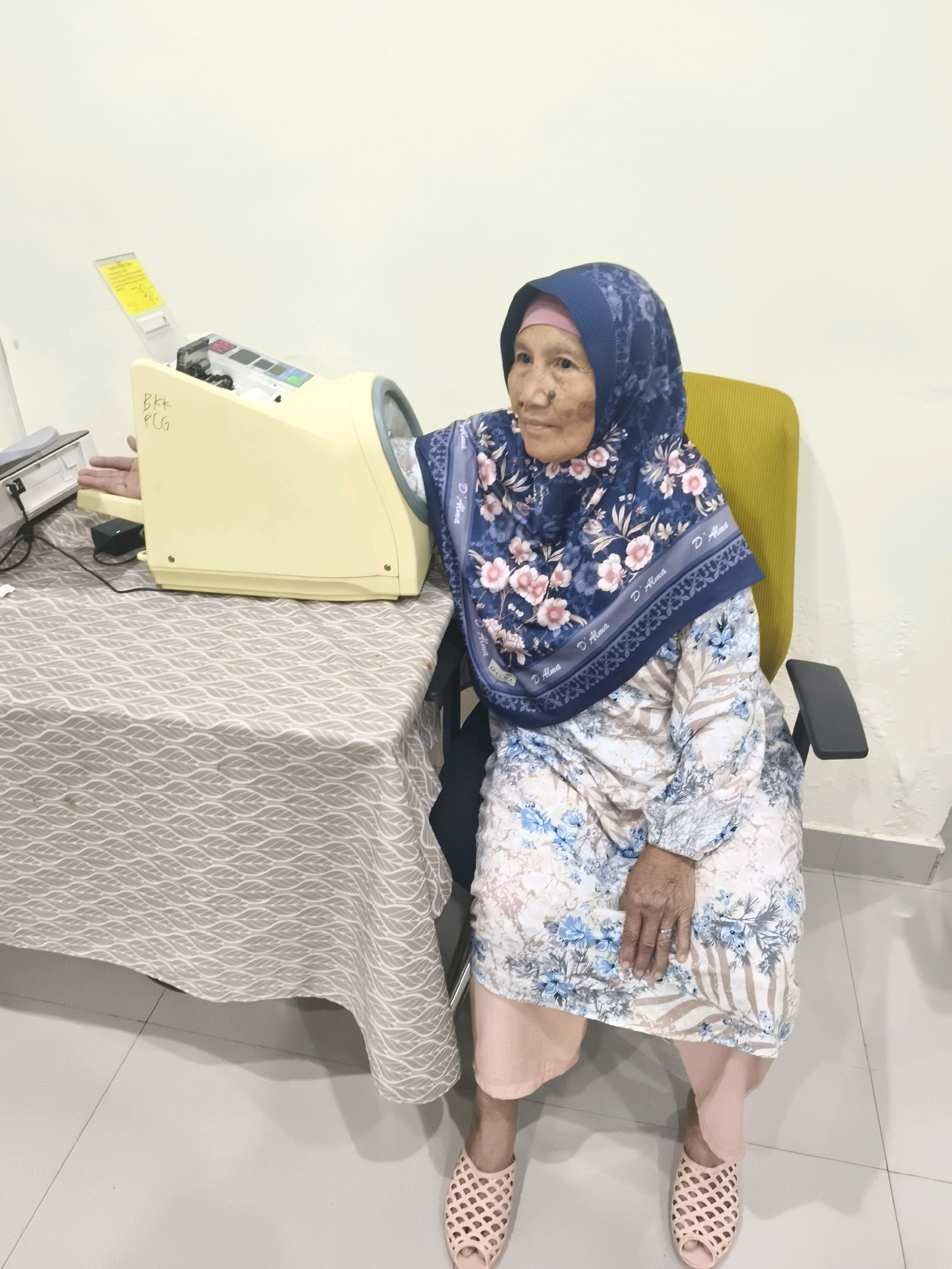 An elderly Indonesian woman, wearing a hijab and a floral-patterned dress, is seated in a yellow office chair next to a table covered with a patterned tablecloth. Her left arm is positioned near a beige medical device, possibly a blood pressure monitor, which has "BKK PCG" written on its side. She is facing to her left, looking away from the camera. The background is a plain, light-colored wall. The scene appears to be indoors, in a clean and well-lit environment, possibly a clinic or a medical facility. The flooring is tiled.
