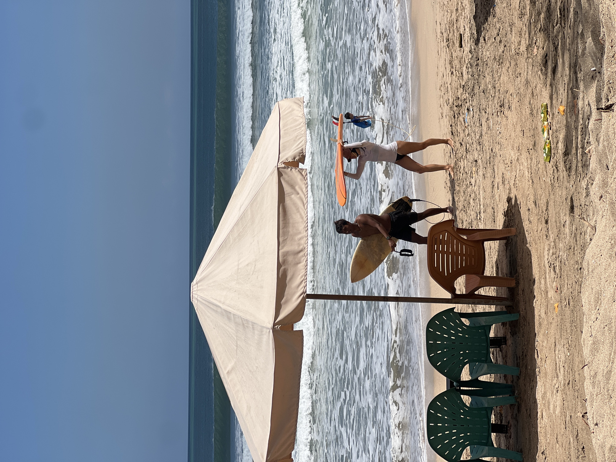 Two individuals are on a sandy beach in Kuta, Indonesia, by the ocean. A large, light-colored umbrella is positioned to the left of the frame, providing shade over three plastic chairs. One chair is brown, and two are green. A man on the left, wearing black shorts, carries a surfboard. A woman on the right, wearing a white long-sleeved top and dark bikini bottoms, carries an orange surfboard above her head. In the background, waves crash on the shore under a clear blue sky.