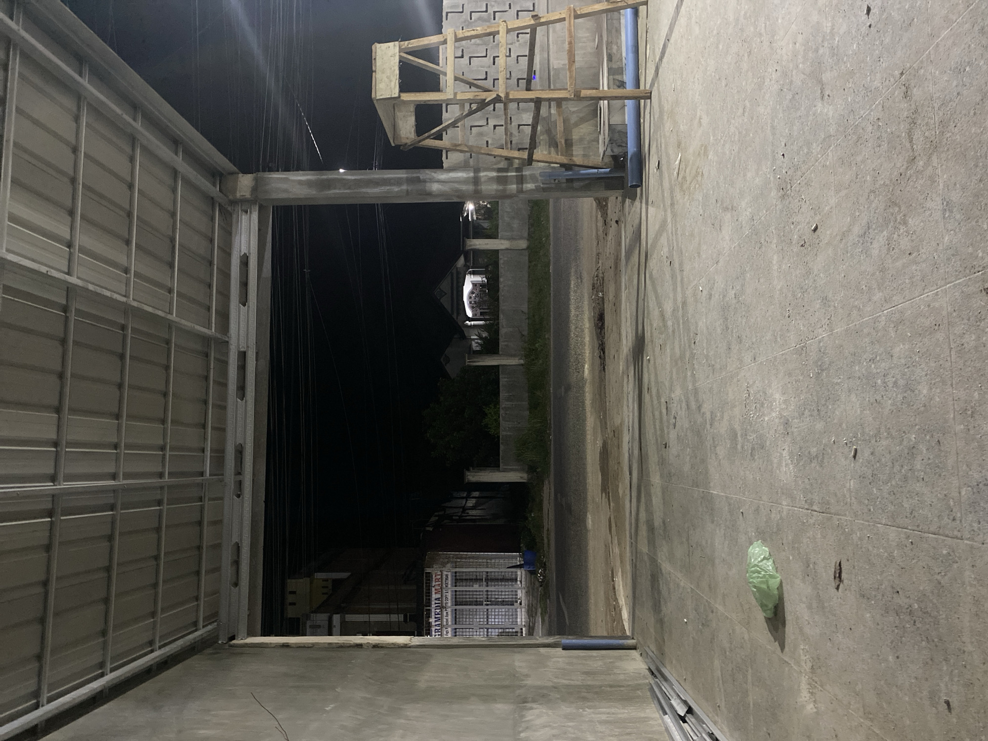 The photo depicts an outdoor scene in Seuneubok Teungoh, Indonesia at night. A metal structure with corrugated panels lines the left side of the frame. A concrete structure occupies the right side. The center reveals a street leading into the darkness. Overhead wires are visible against the night sky. Further down the street, buildings with lights can be seen. A wooden frame structure sits atop the concrete structure near the foreground. A green plastic bag is visible on the concrete structure. A partially visible sign for "SRAMBINA MART" is present in the distance.