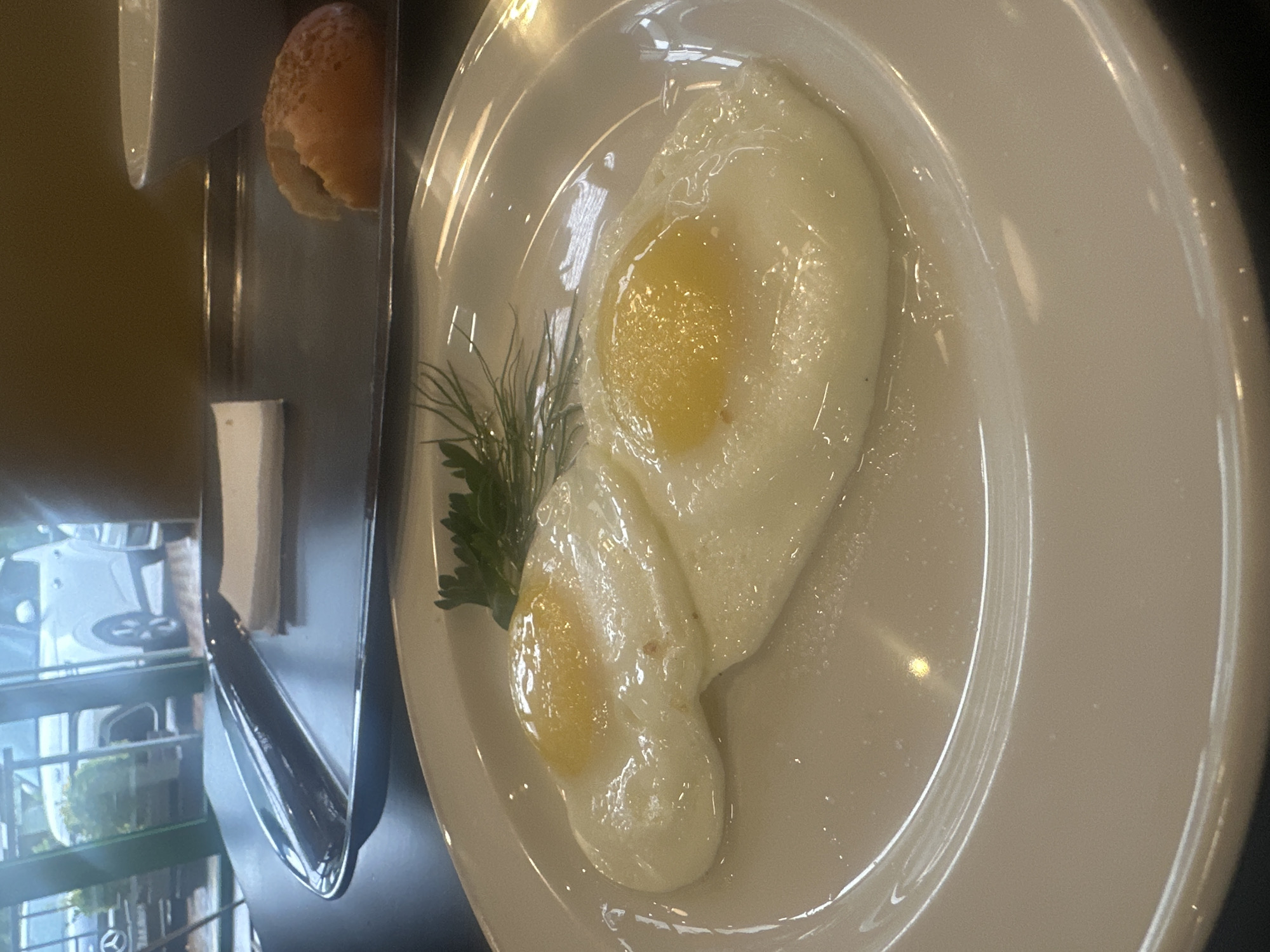 The image captures a meal presentation likely photographed in Krasnaya Polyana, Russia, given the presence of a reflection of a Mercedes-Benz dealership. The focal point is a white plate featuring two fried eggs. Garnish includes a small sprig of dill and parsley. Adjacent to the plate is a narrow metal tray containing a small white container, a single bun, and what appears to be a pat of butter or cheese. The scene appears to be inside a restaurant or cafe due to the reflective glass and likely outdoor environment.
