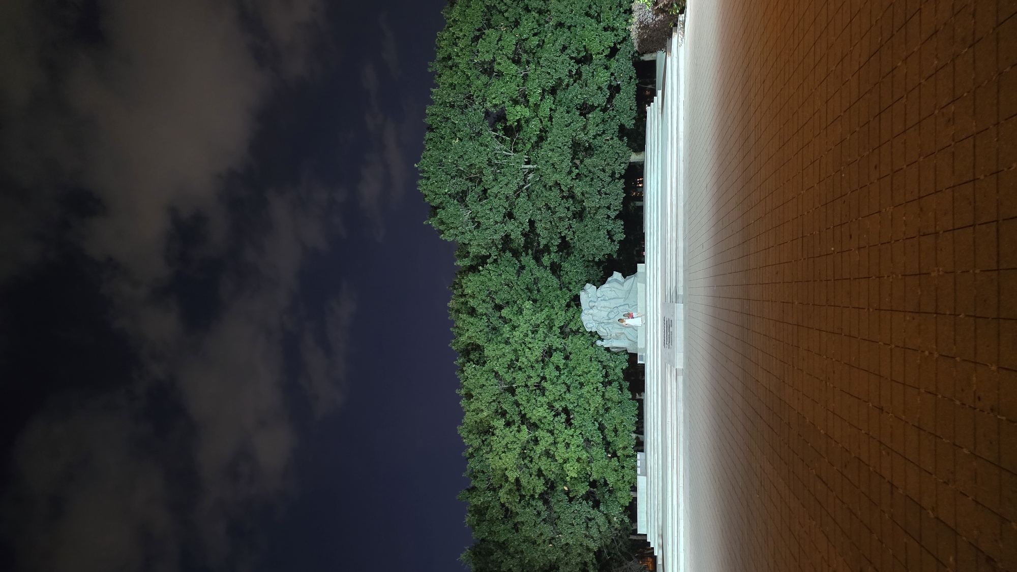 The image depicts a monument and a row of trees at night. Two individuals, one adult and one child, are standing near the base of the monument. The monument is situated on a multi-tiered platform, and a paved area extends towards the viewer. The sky above is dark with scattered clouds. The monument and trees are illuminated by artificial light, casting shadows on the paved ground. The overall scene appears to be a public space, possibly a park or memorial site. This scene is located in Krasnodar, Russia.