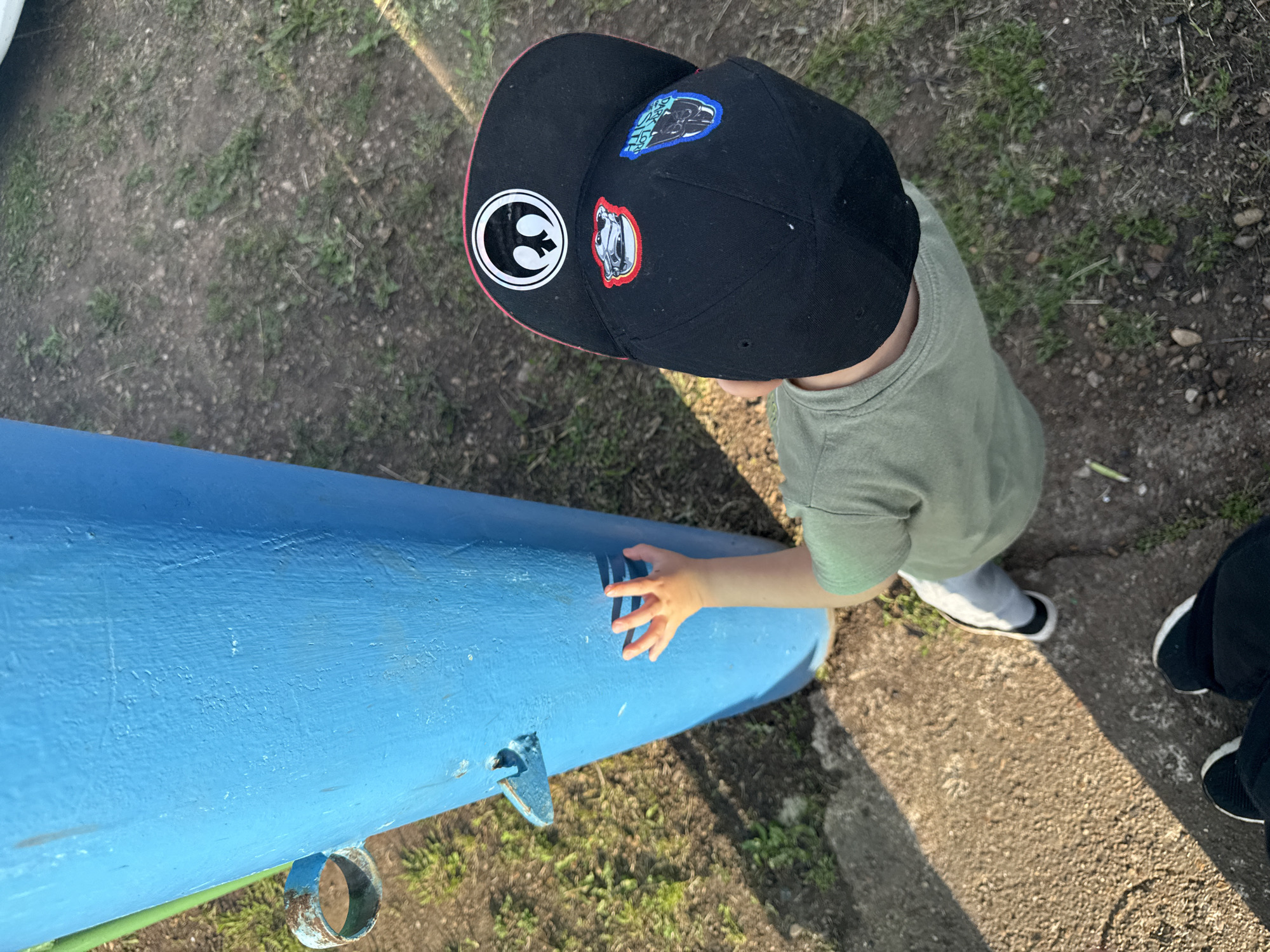 A young child wearing a green t-shirt and a black baseball cap with patches is reaching out to touch a bright blue metal pole in an outdoor setting. The child's left hand is extended, fingers spread, towards the pole. The ground around the pole is a mix of dirt and sparse grass. In the foreground, two pairs of feet are visible; one pair belongs to the child, wearing black and white sneakers, and the other pair, in black shoes, suggests an adult is present, possibly taking the photo. The lighting indicates it is daytime, with shadows cast across the ground. The location is identified as Чуй-Атасево, Russia.