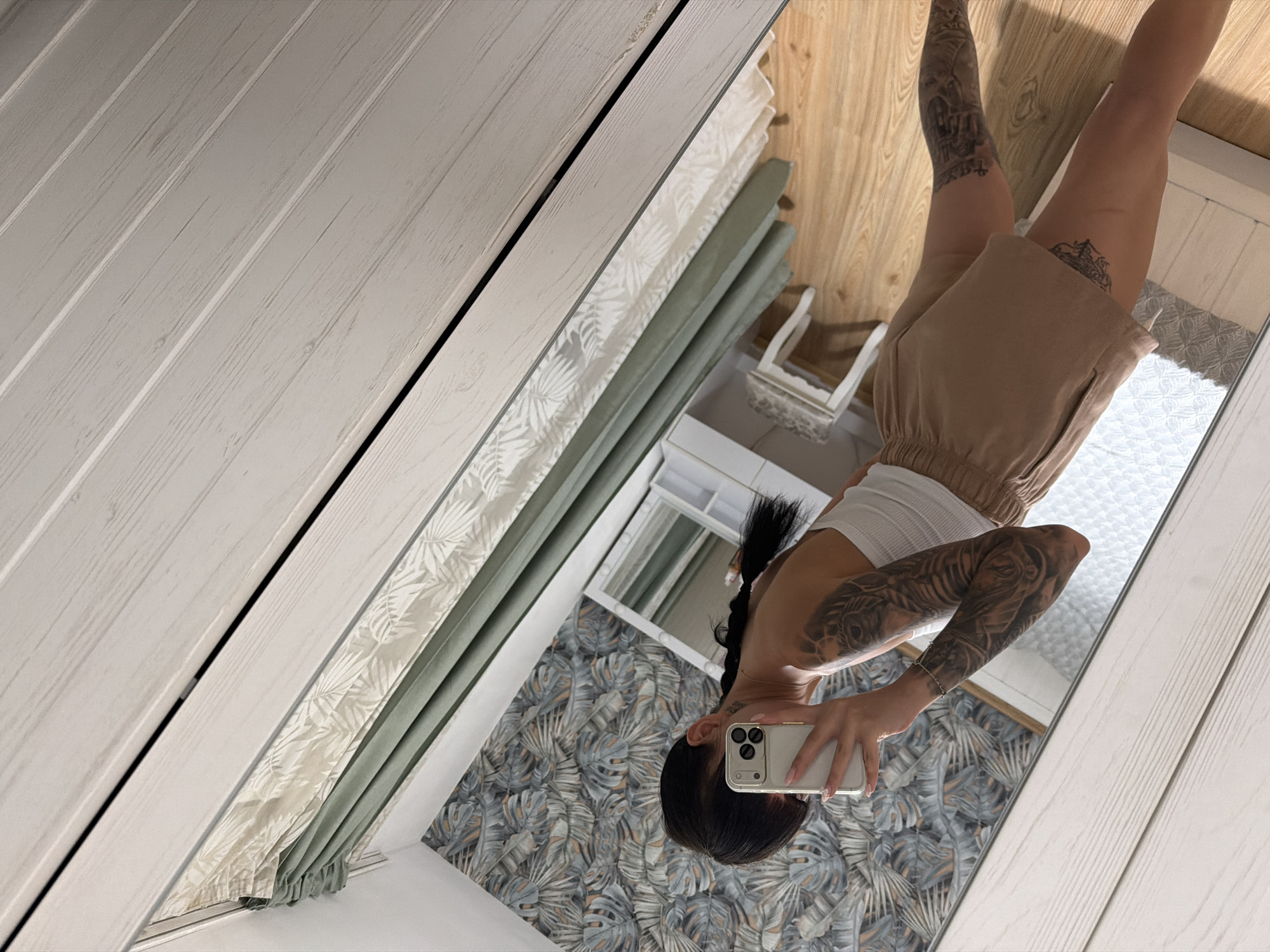 The image shows a young woman taking a mirror selfie in what appears to be a bedroom. She has dark hair styled in a braid and is wearing a white tank top and tan shorts. Her arms and legs are covered in extensive tattoos. In the reflection, we see a dresser with a mirror, a small stool, and a wall with tropical-themed wallpaper. The room has wooden floors and is decorated with curtains. The image itself is taken from a high angle, with the camera tilted. There is no visible indication of the location being in Plovdiv, Bulgaria.